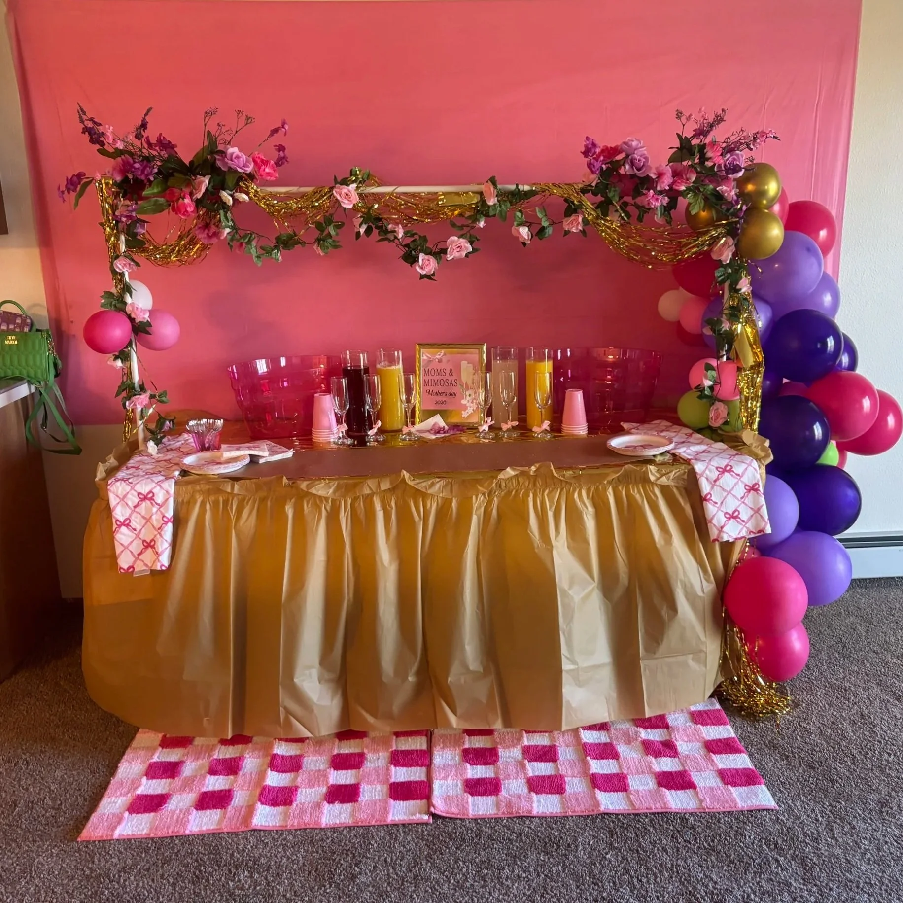 Decorative table setup for Mother's Day with pink, purple, and gold balloons on a gold table skirt, floral garlands, pink papel picado table runner, and a pink backdrop. Drinks, plates, and a framed sign reading "Moms & Mimosas Mothers Day 2020" are on the table.