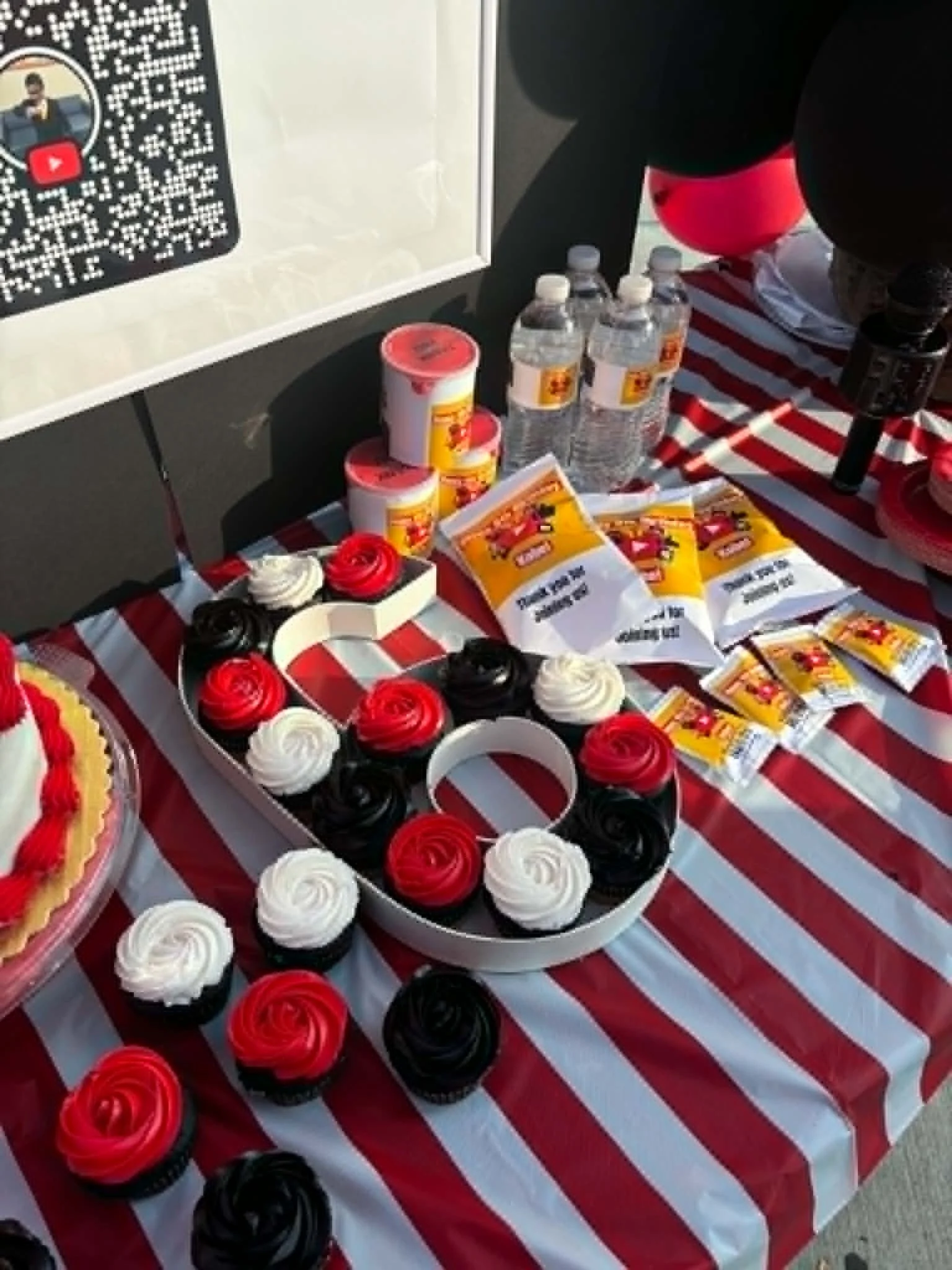 Table with cupcakes decorated as roses in red, white, and black, and bottled water with a QR code sign.