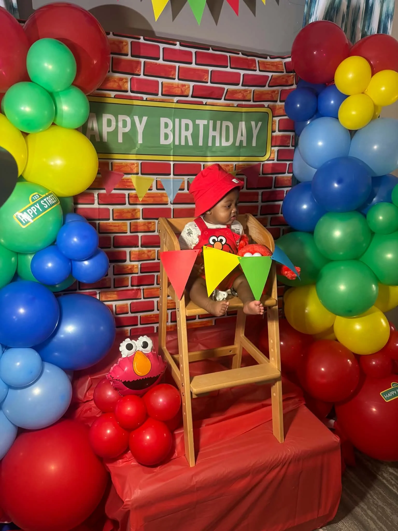 Child sitting in wooden high chair surrounded by colorful balloons in red, blue, green, yellow, and black. The background has a brick wall design with a green sign reading 'HAPPY BIRTHDAY' and multicolored pennant banners, indicating a birthday celebration.