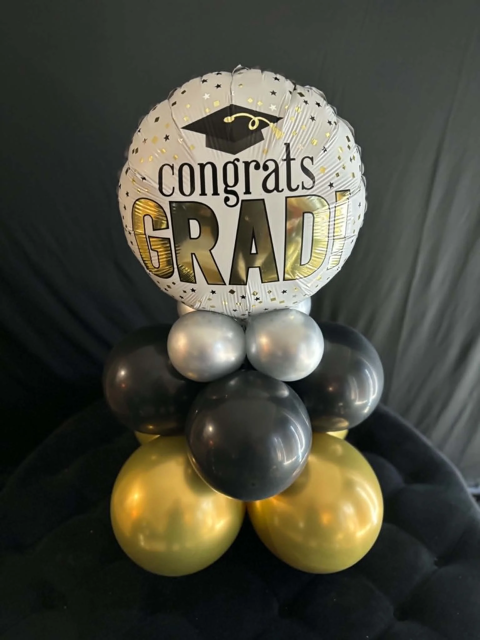 Balloon arrangement with a large round foil balloon that says 'congrats GRAD' with a graduation cap icon, surrounded by smaller gold, silver, black, and yellow balloons.