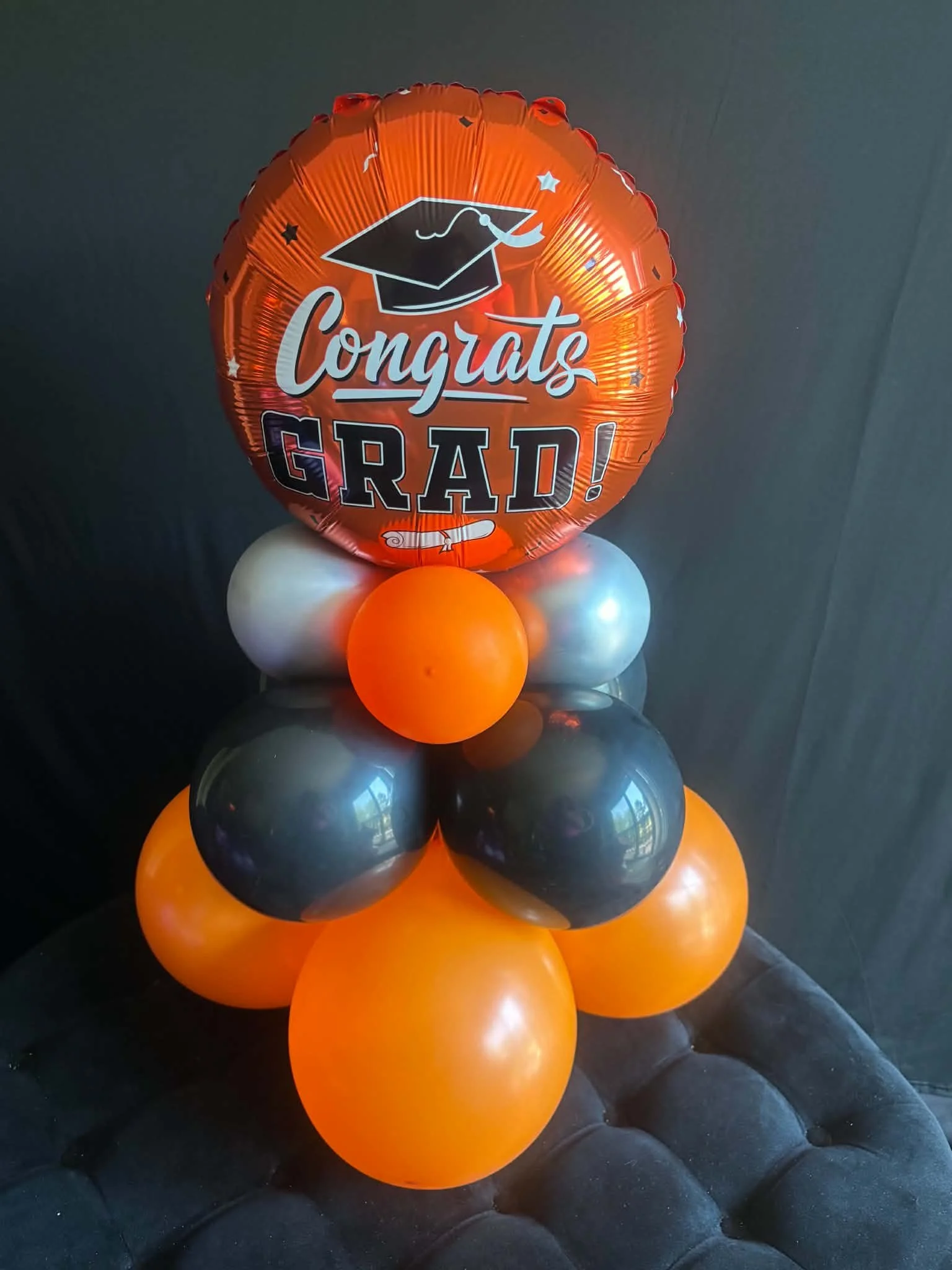 A balloon arrangement with a large round orange balloon that has a graduation-themed message, including a black graduation cap illustration and the words 'Congrats Grad!' on it. The large balloon is surrounded by smaller orange, black, and silver balloons.