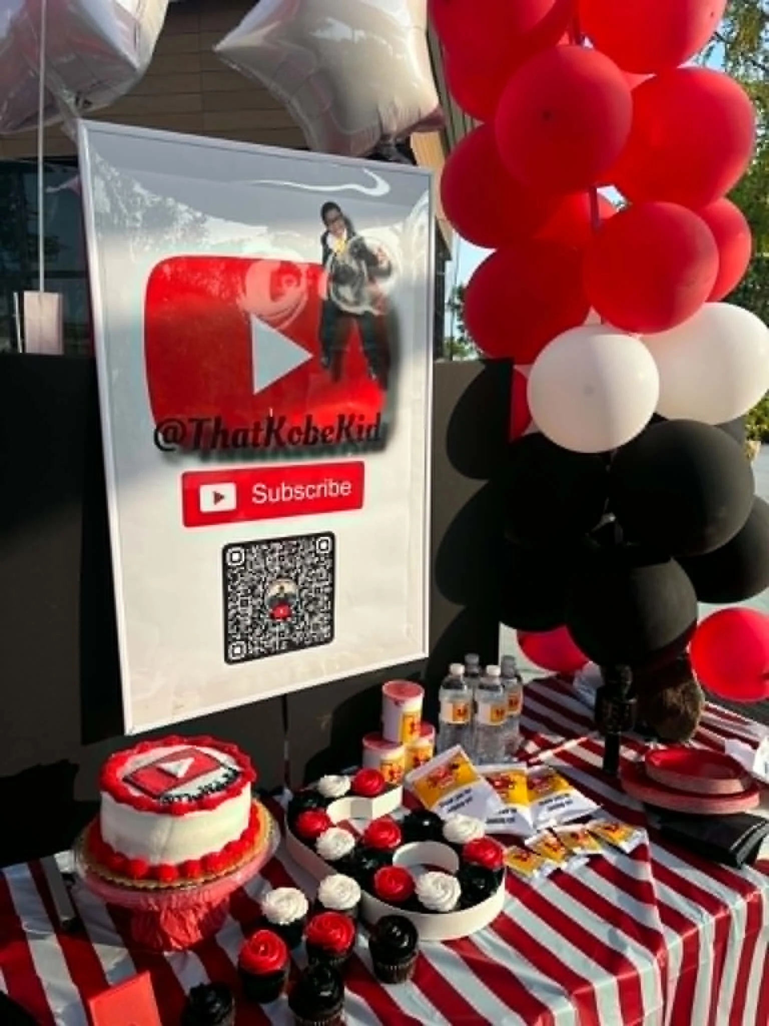 Table decorated with red, black, and white items, including cupcakes, balloons, gifts, and a large YouTube-themed cake, at an outdoor event.