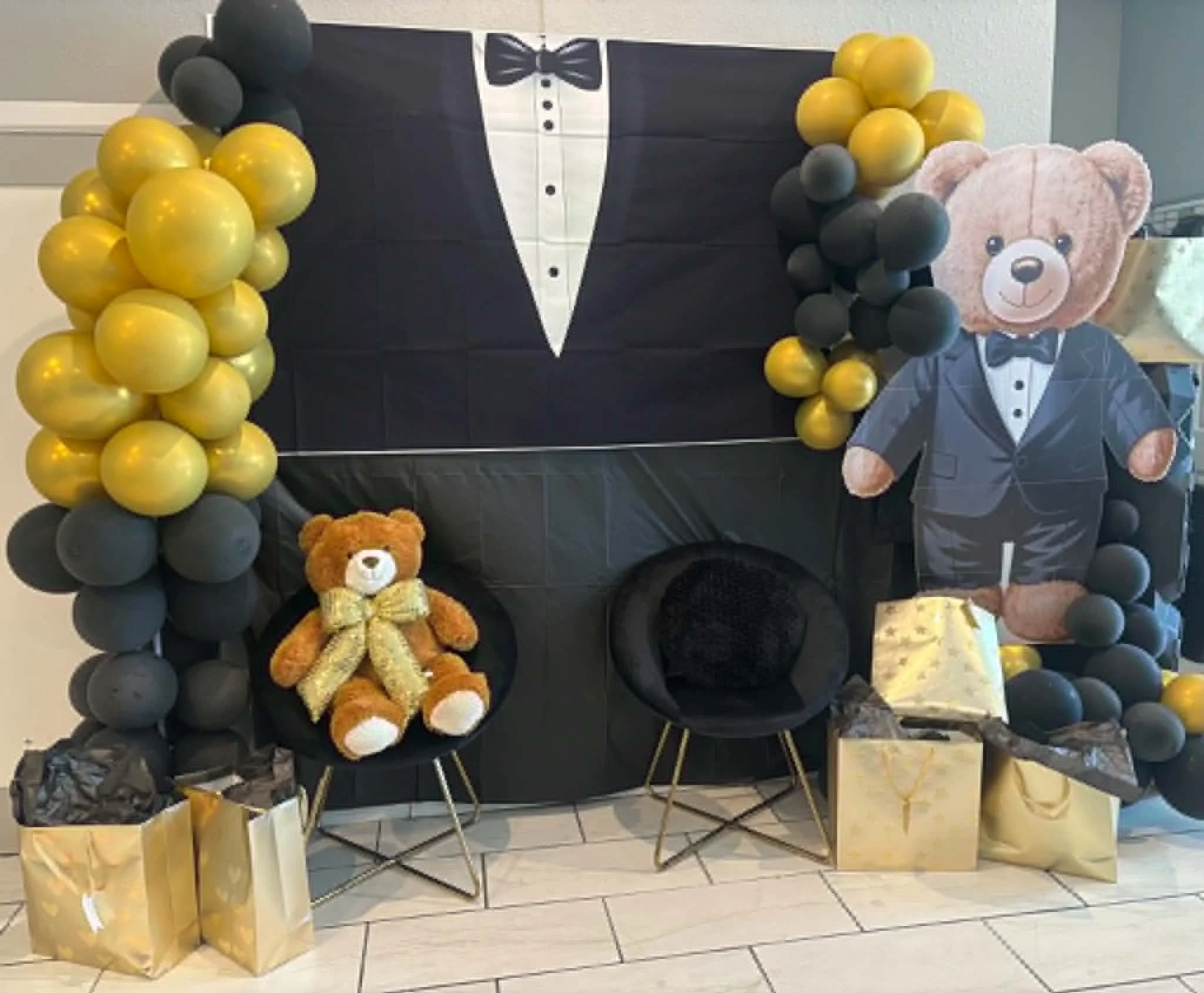 Decorative display with a large black and white backdrop resembling a tuxedo, accented with yellow and black balloons arranged around it. A cardboard cutout of a bear in a tuxedo poses in front, complemented by plush teddy bears and yellow gift bags on the floor.