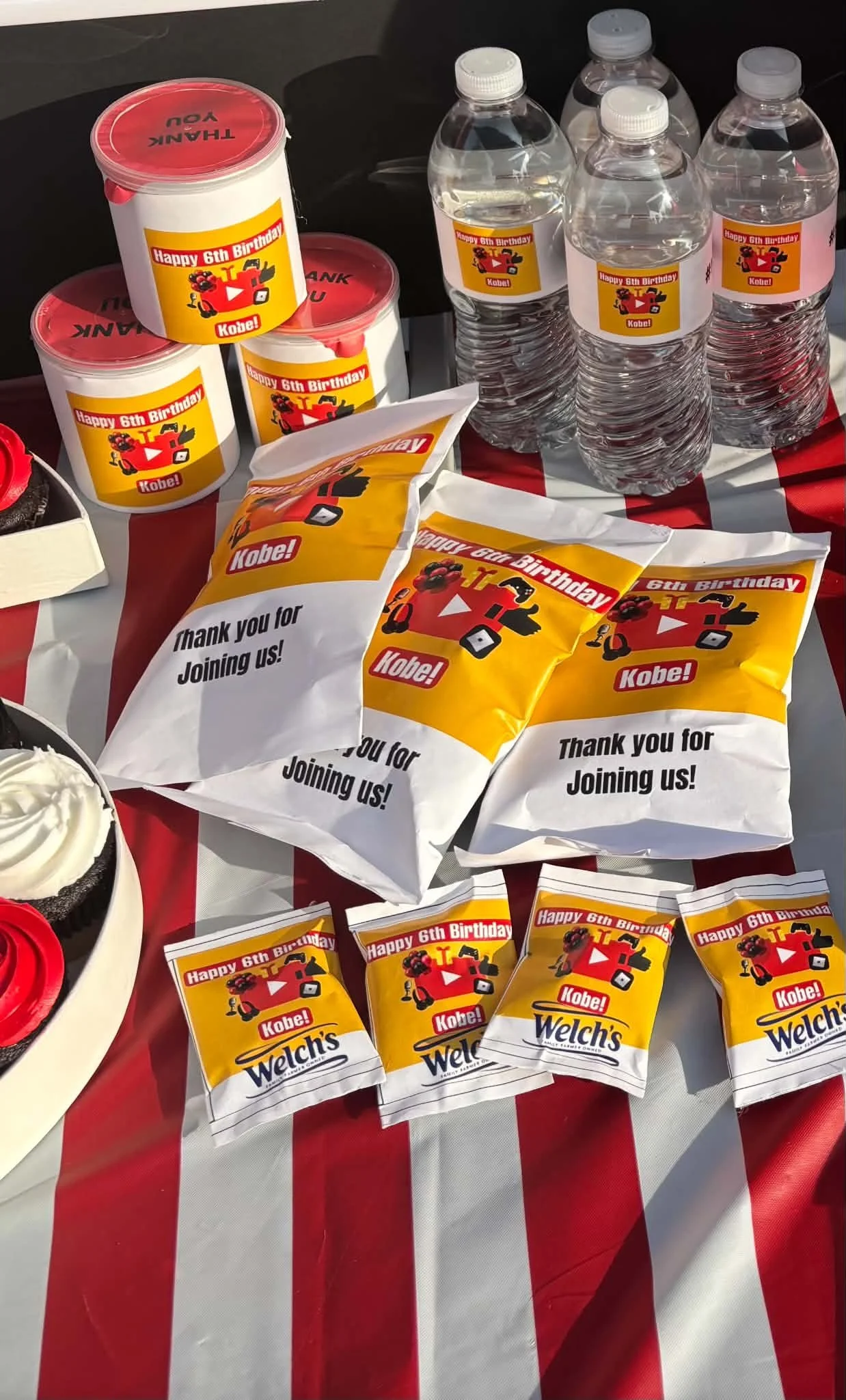 Party table decorated with red and white striped tablecloth, featuring bottled water with birthday labels, snacks, and cupcakes with red frosting.