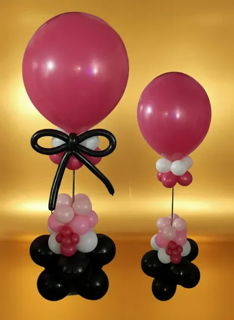 Two balloon sculptures with large pink balloons, a variety of smaller balloons in pink, white, and black, and balloon arrangements resembling flower bases, set against a golden background.