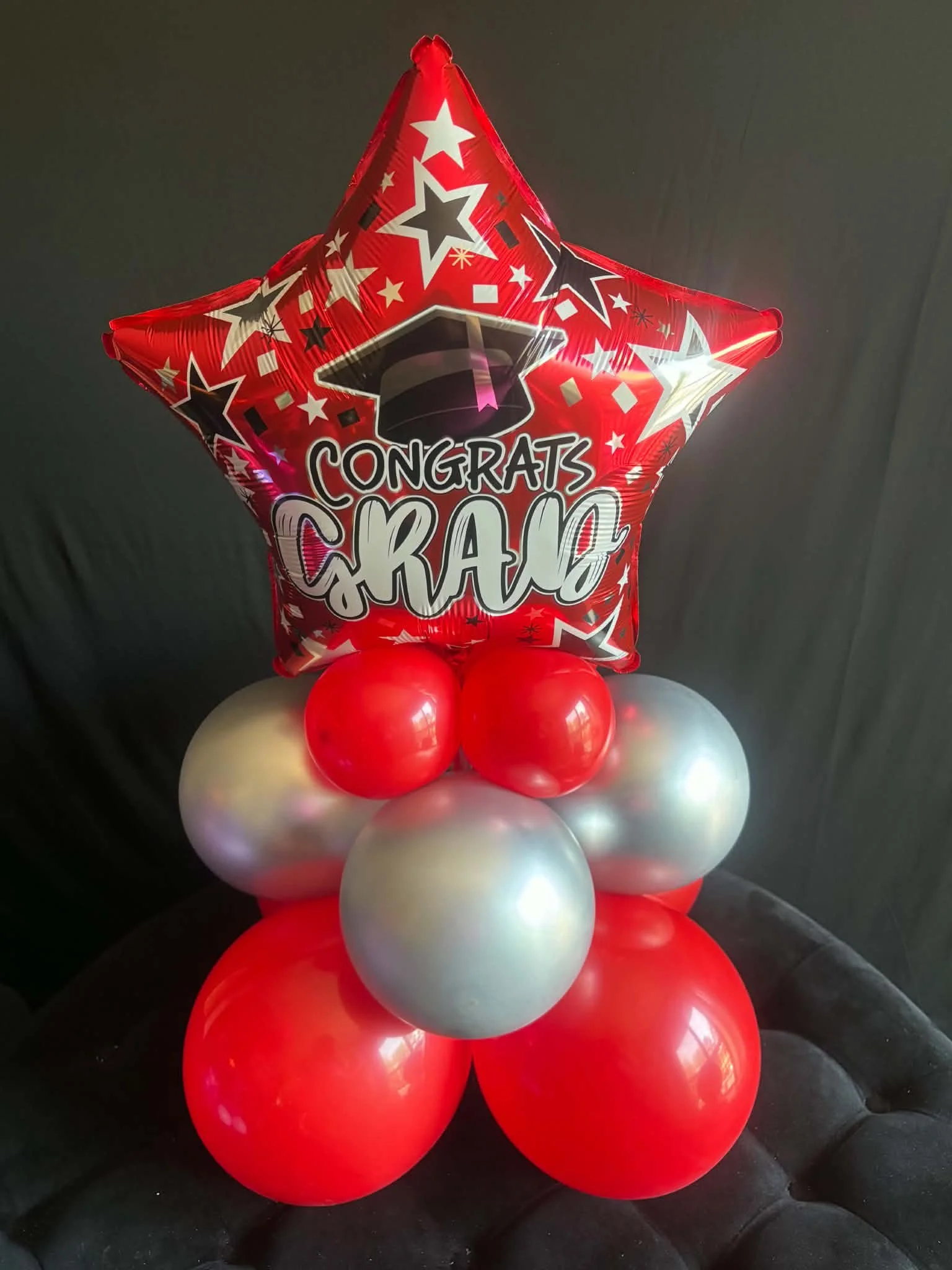 A cluster of red, white, and silver balloons arranged for a celebration, with a large red star-shaped balloon on top that has a message saying 'Congrats Grad' with a graduation cap design.
