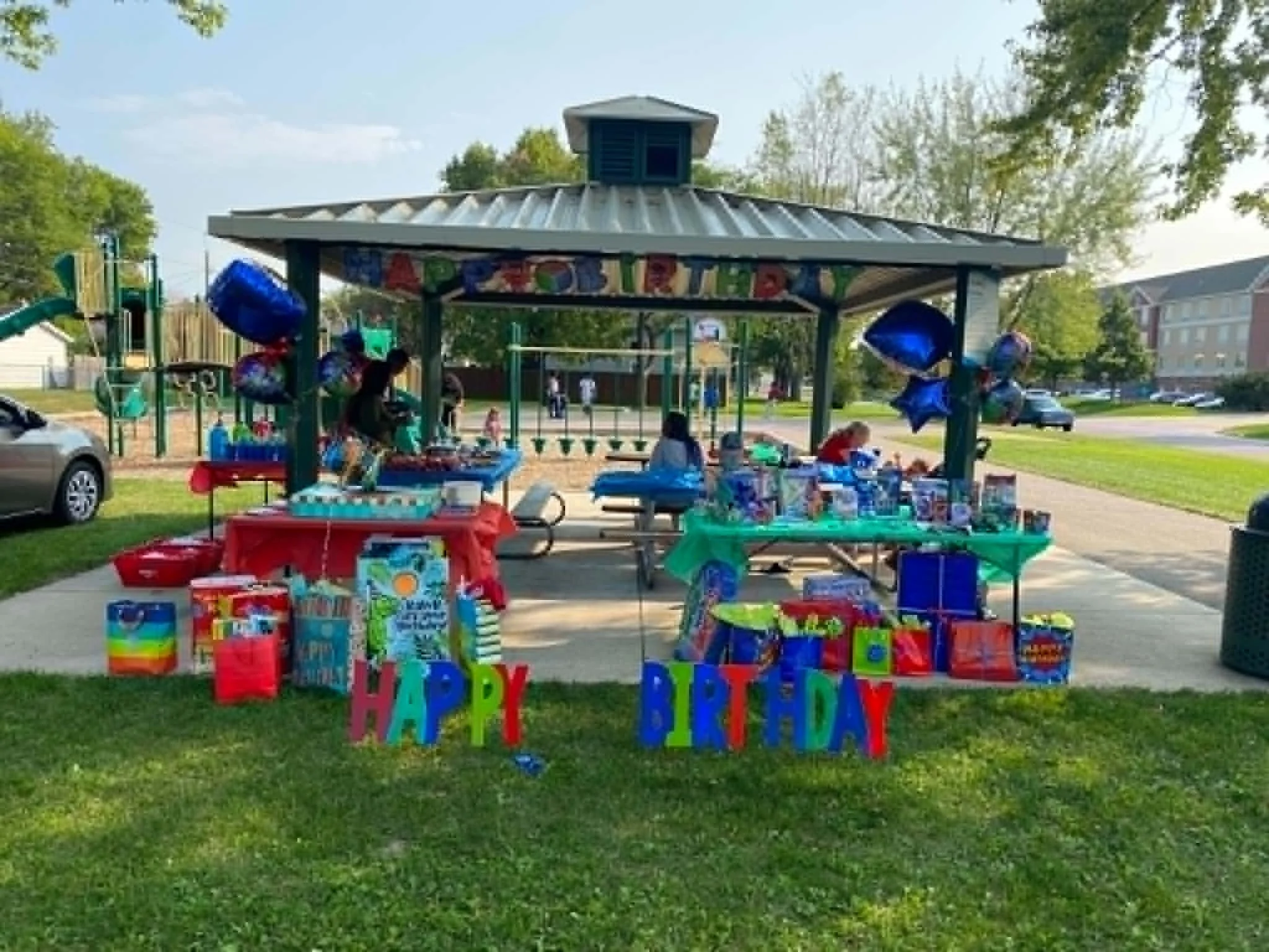 Outdoor birthday party setup with a colorful table, balloons, and a 'Happy Birthday' banner under a gazebo in a park.