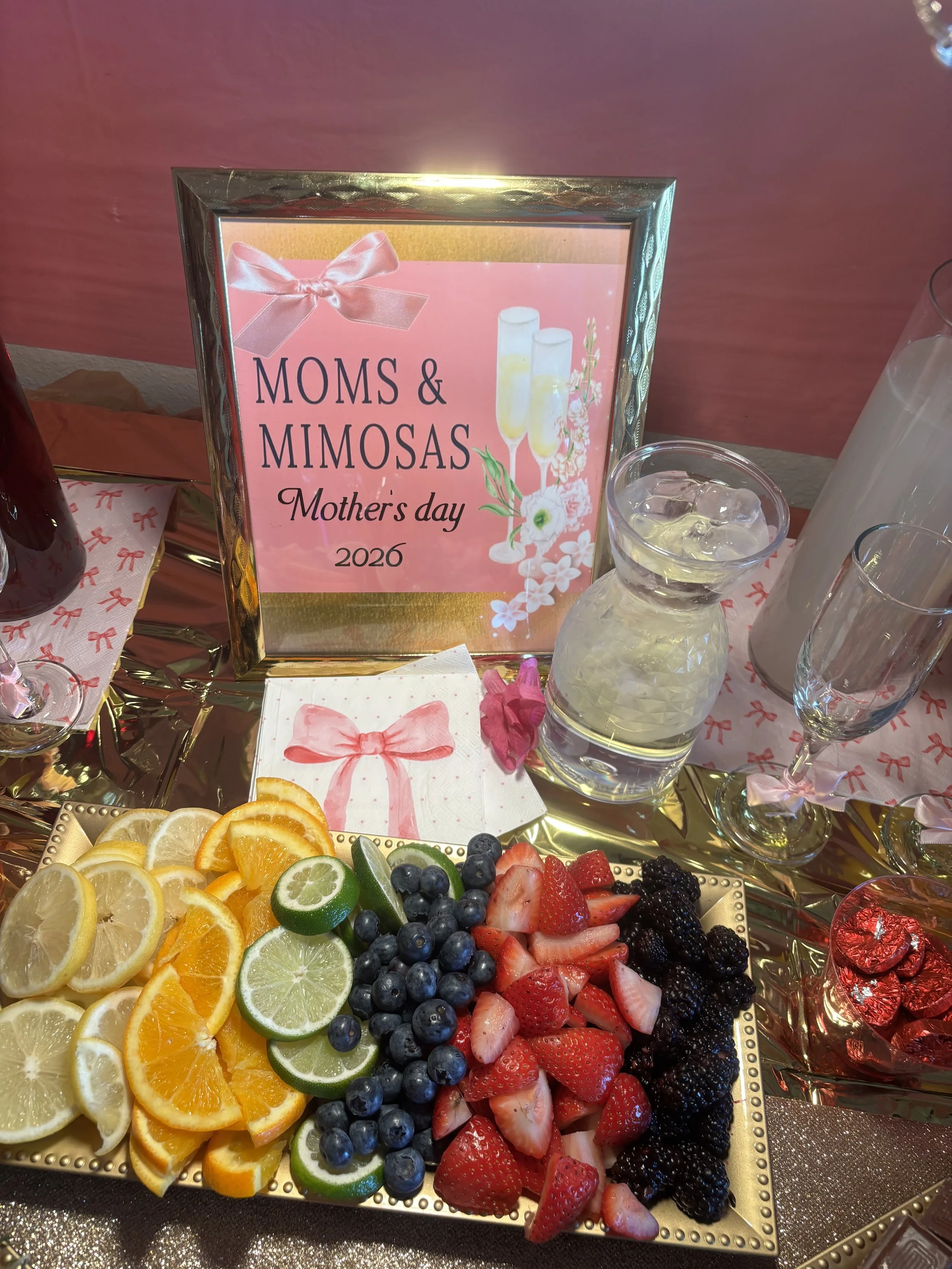 A decorative tray of sliced citrus fruits including lemon, lime, orange, and lemon. The tray is surrounded by other glassware and a framed sign that reads "MOMS & MIMOSAS, Mother's Day 2026."