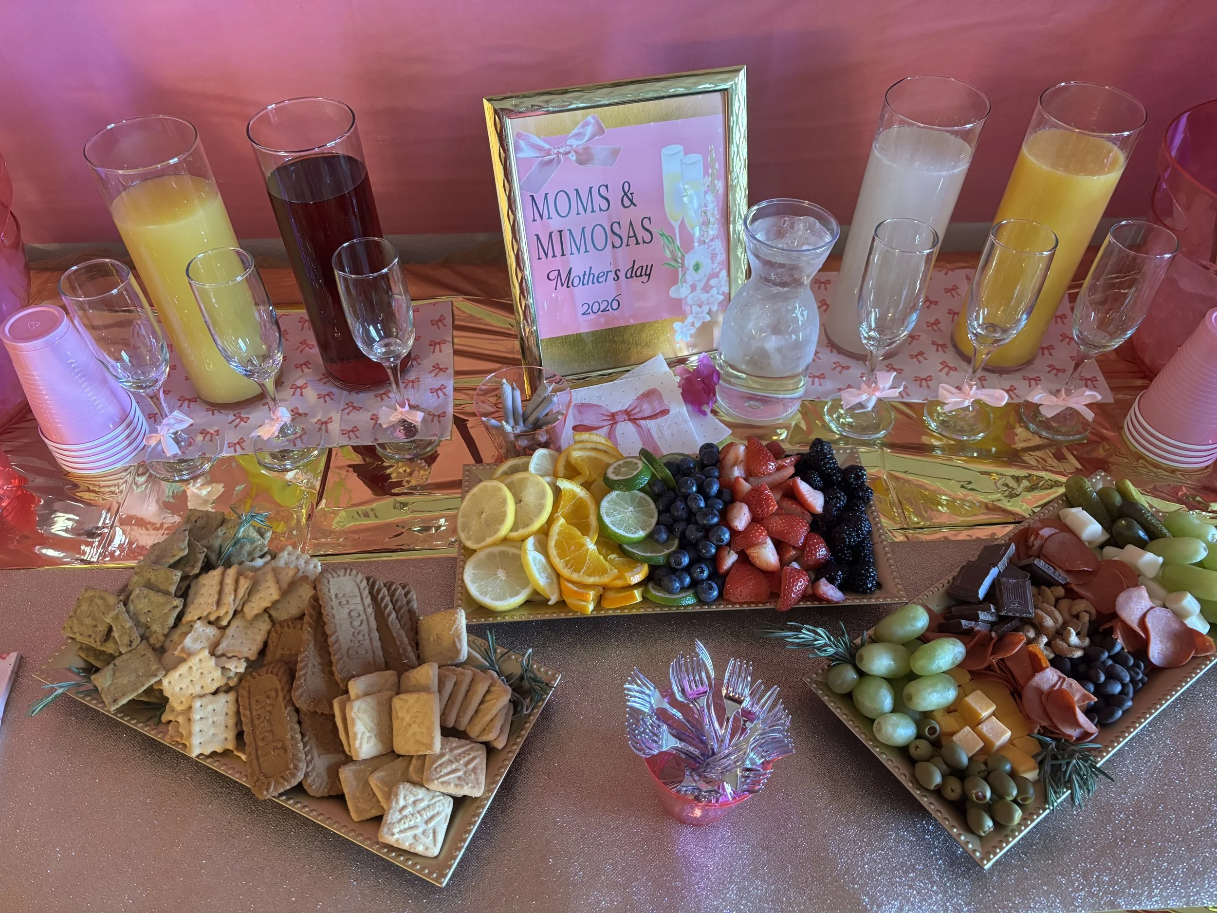 Decorative table setting for Mother's Day with trays of crackers, cookies, and cheese, along with bowls of fresh fruits and vegetables, champagne flutes, and a framed sign that reads "Moms & Mimosas Mother's Day 2026".