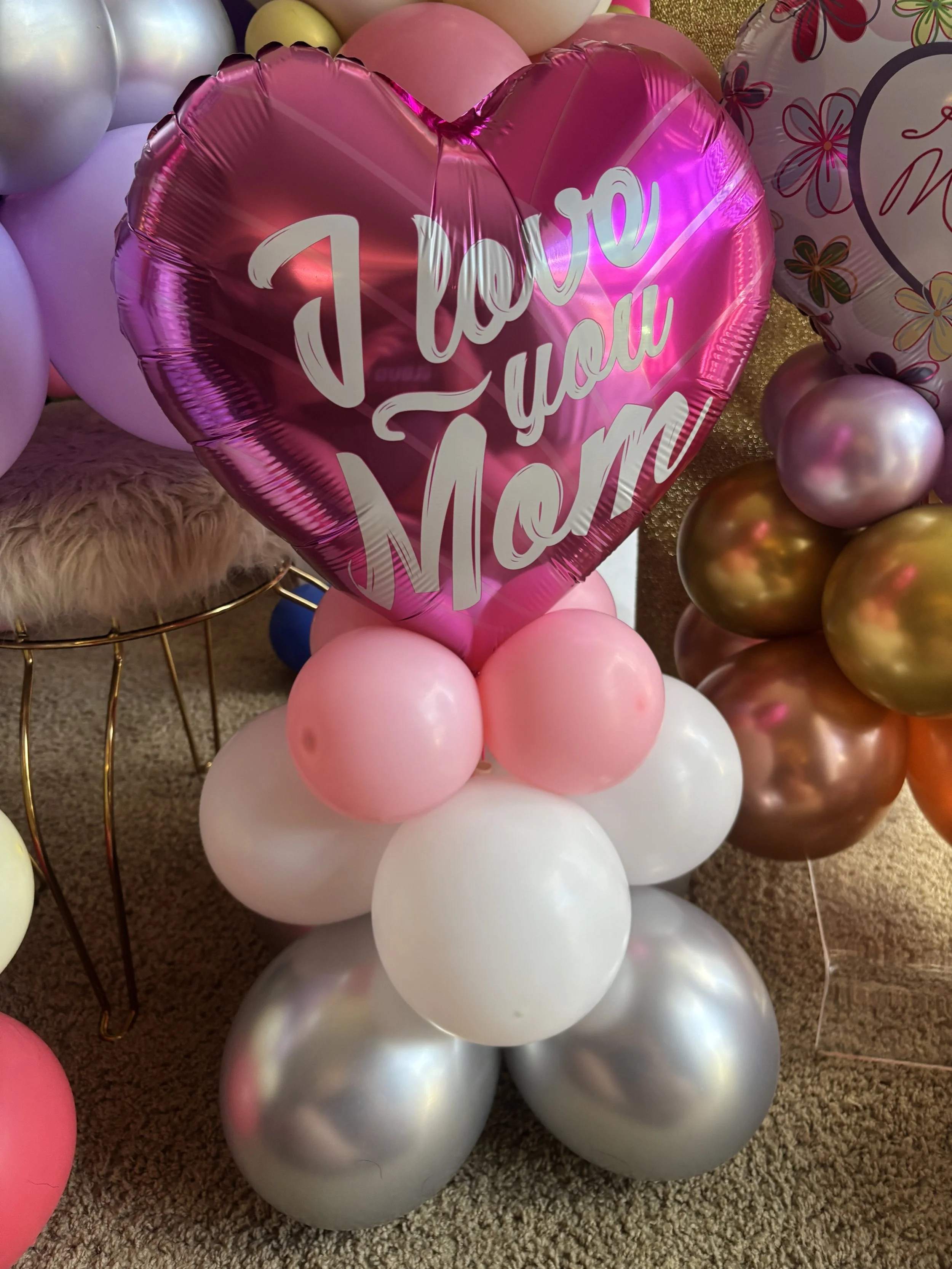 Bunch of pink, white, gold, and silver balloons, including a large pink heart-shaped balloon with the words 'I love you Mom' written on it.