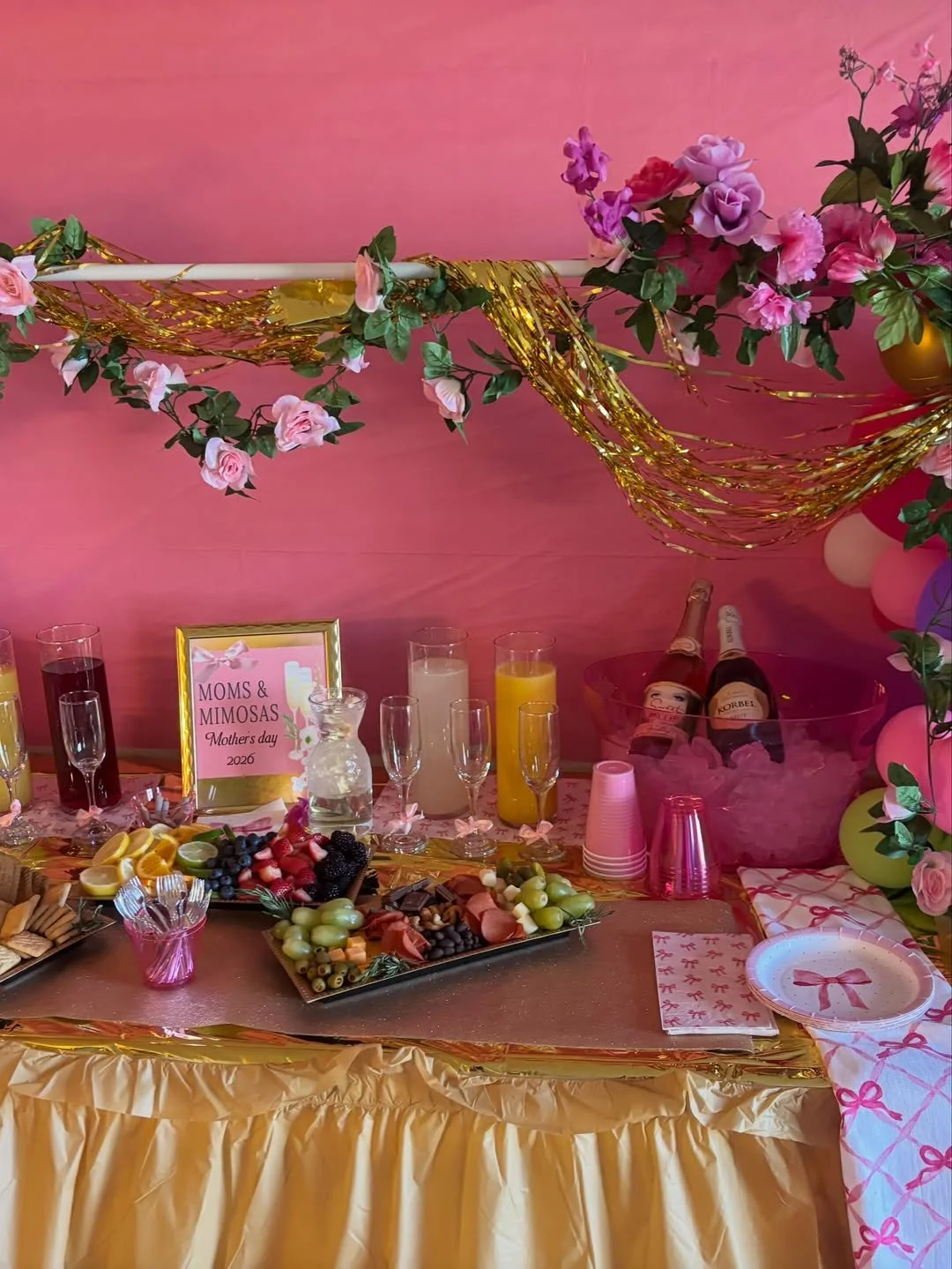 Decorated Mother's Day celebration table with pink and gold accents, flowers, fruit, drinks, and gifts.