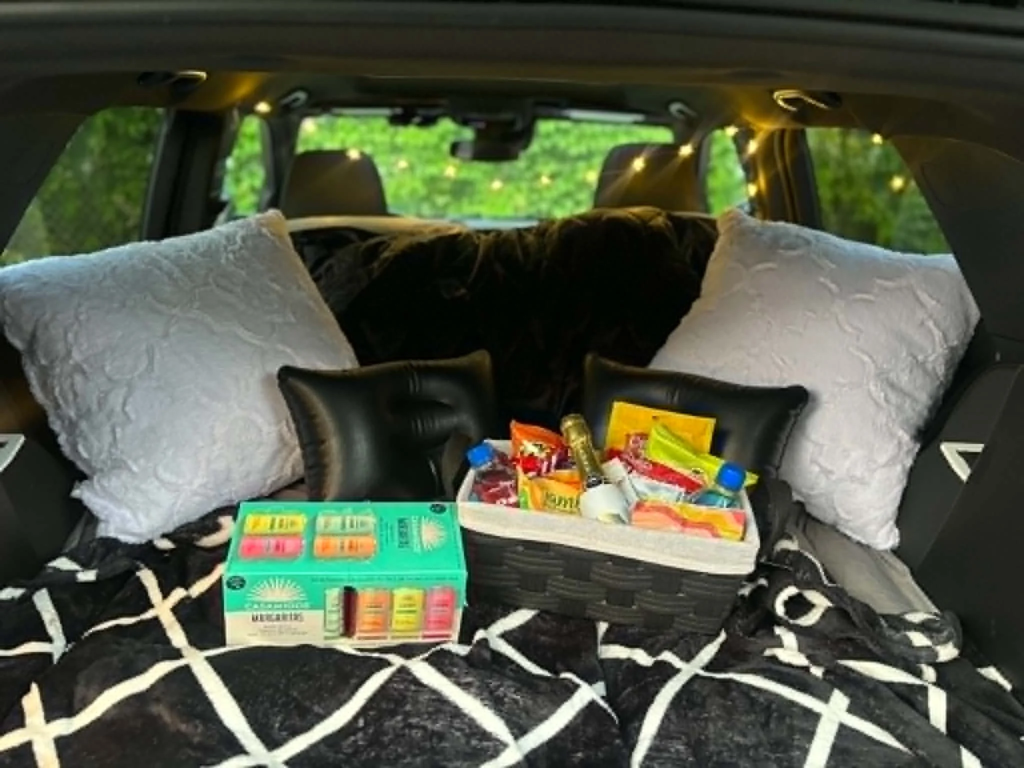 Backseat of a vehicle filled with various items including a white pillow, snacks, and colorful candy, with a green outdoor scene visible through the windows.