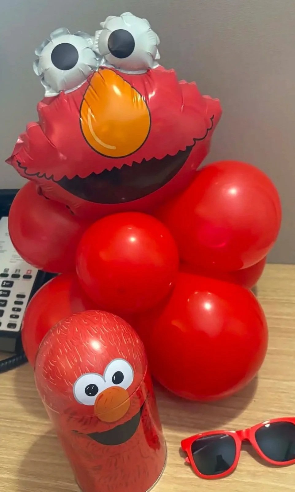 A cluster of red balloons with a large Elmo face balloon on top and an Elmo-themed water bottle, alongside red sunglasses on a wooden surface.