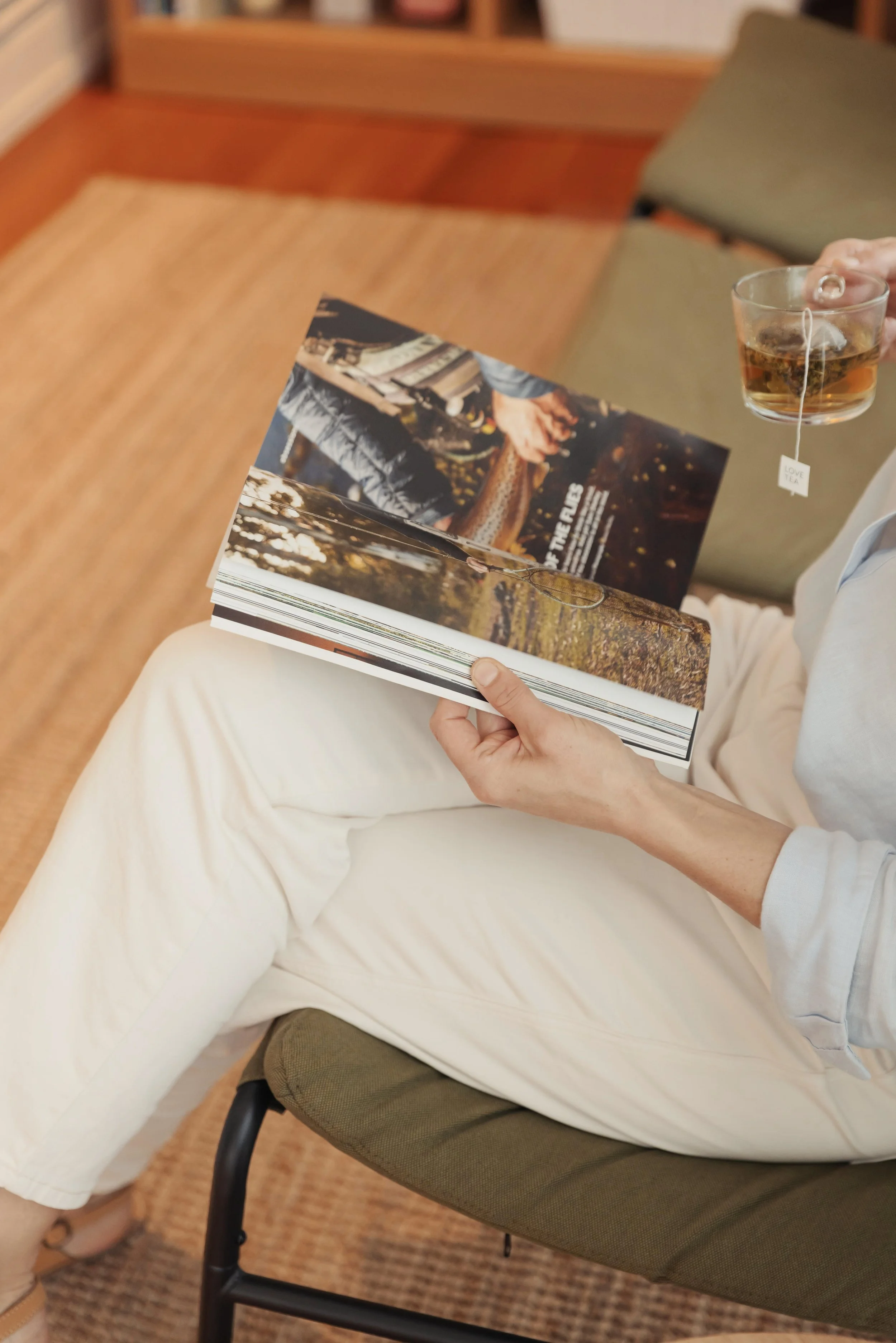 A woman sitting on a chair in a somatic nutrition therapy clinic waiting room, holding an open magazine in one hand and a glass of tea in the other.