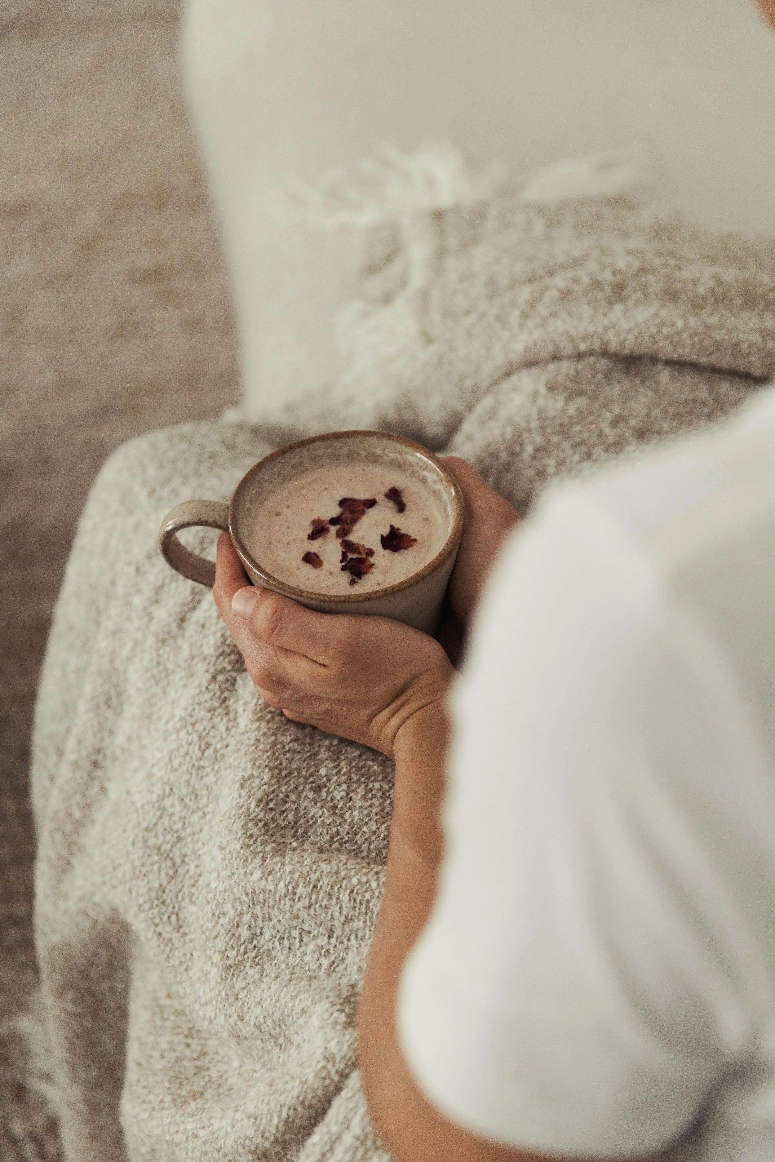 A woman sitting on a couch holding a mug of hot cacao topped with rose petals, with a soft, beige blanket over her lap.