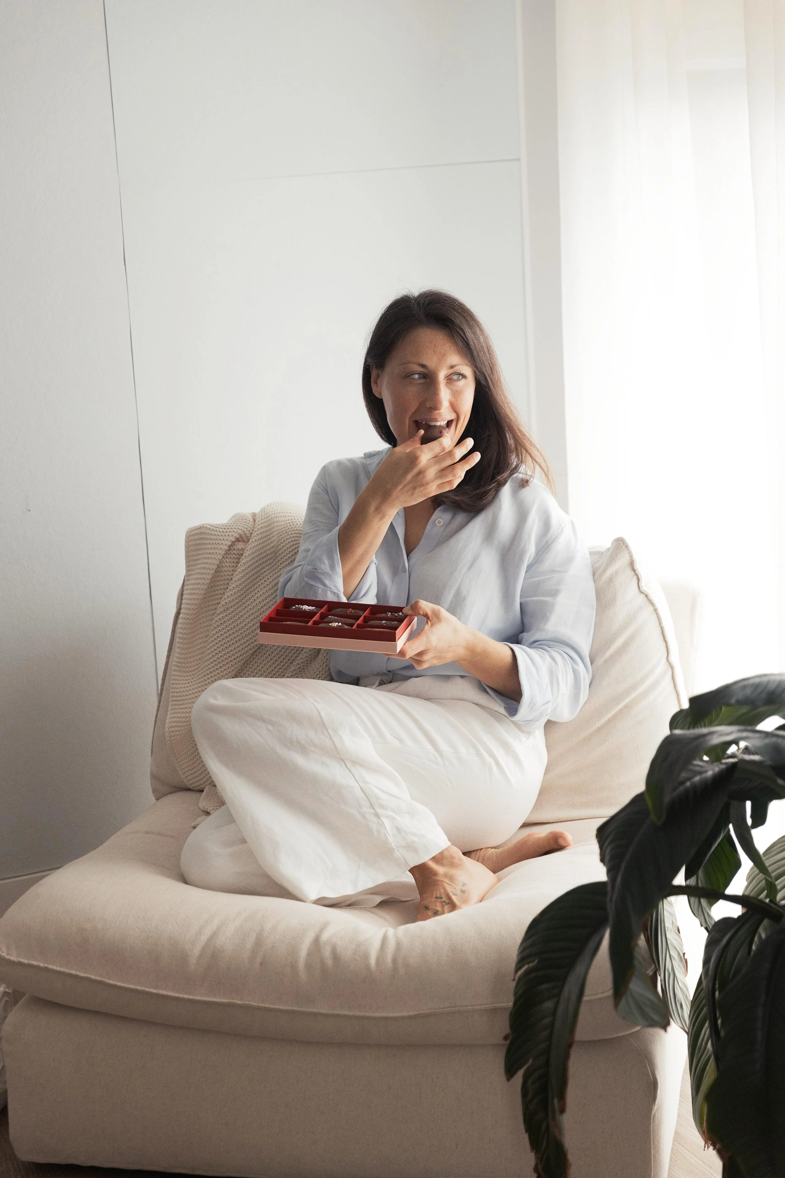 Somatic nutrition therapist and dietitian Emily Hahn sitting on a beige couch, holding a box of chocolates, smiling and tasting one, with a plant in the foreground and a bright window in the background.