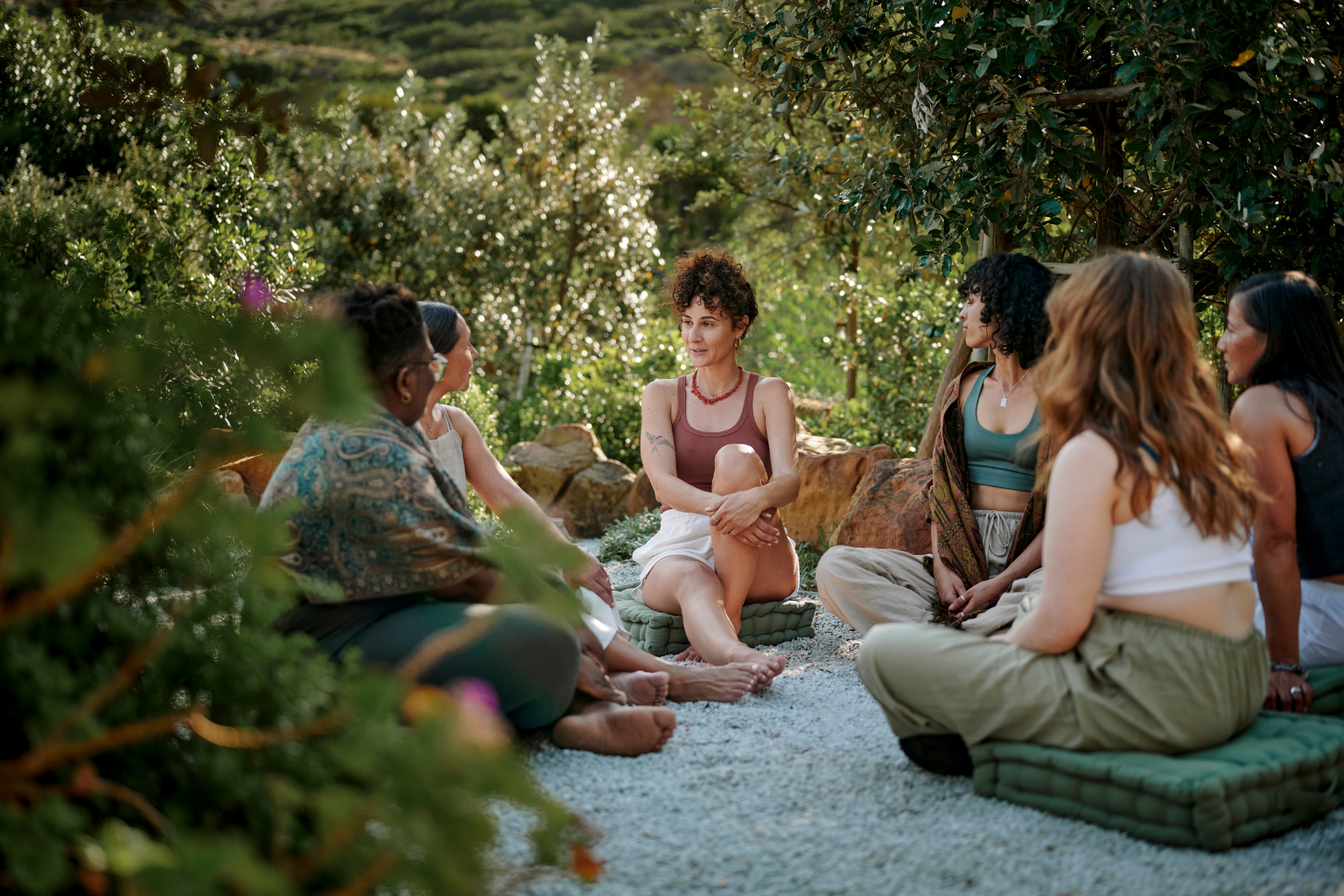 A diverse group of 6 women, of varying ethnicities and body sizes, sitting in a women's circle outdoors on green cushions, connecting through conversation and smiling, surrounded by greenery and trees.