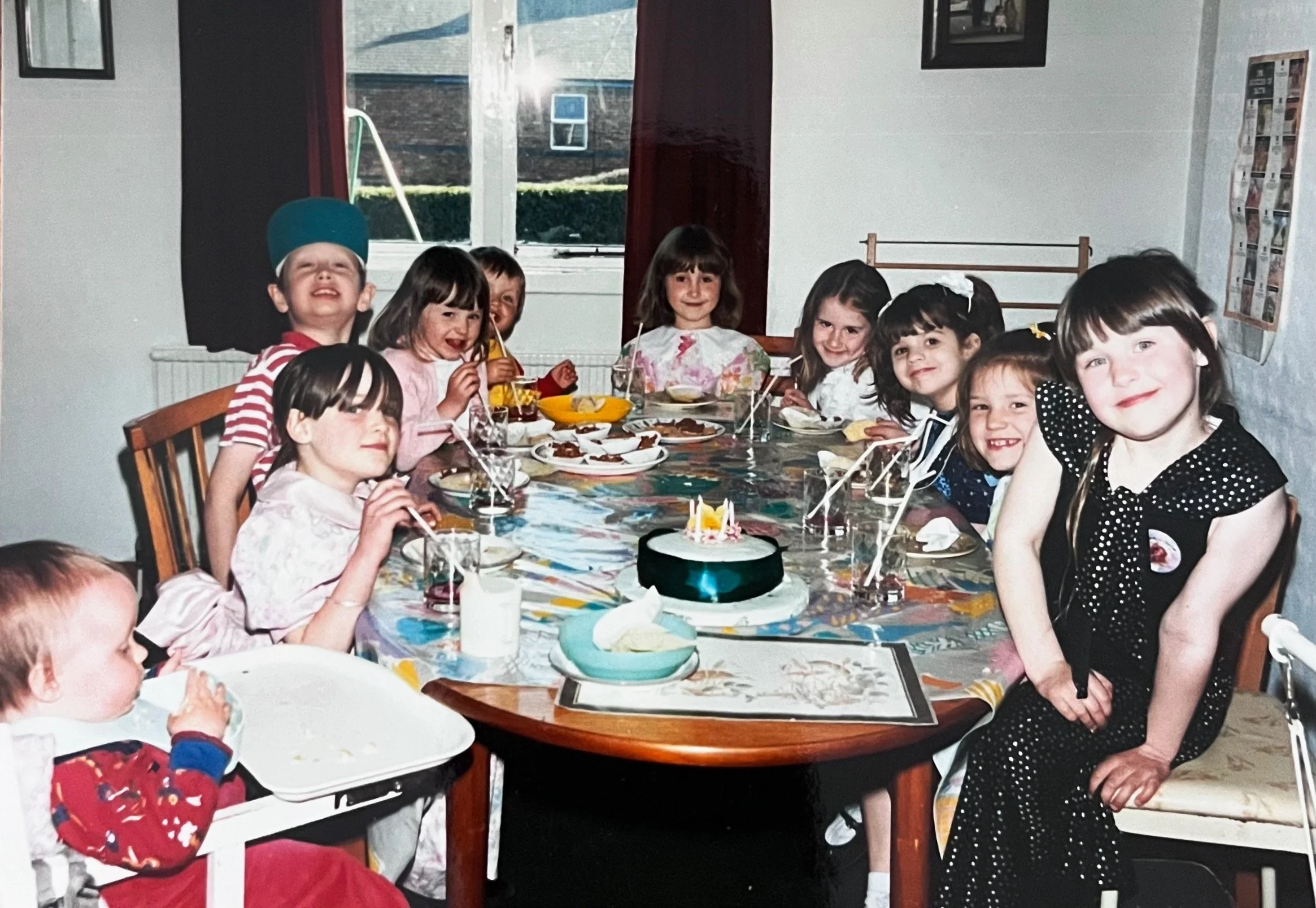 Children gathered around a birthday cake at a party, celebrating with party hats, drinks, and desserts in a decorated room.