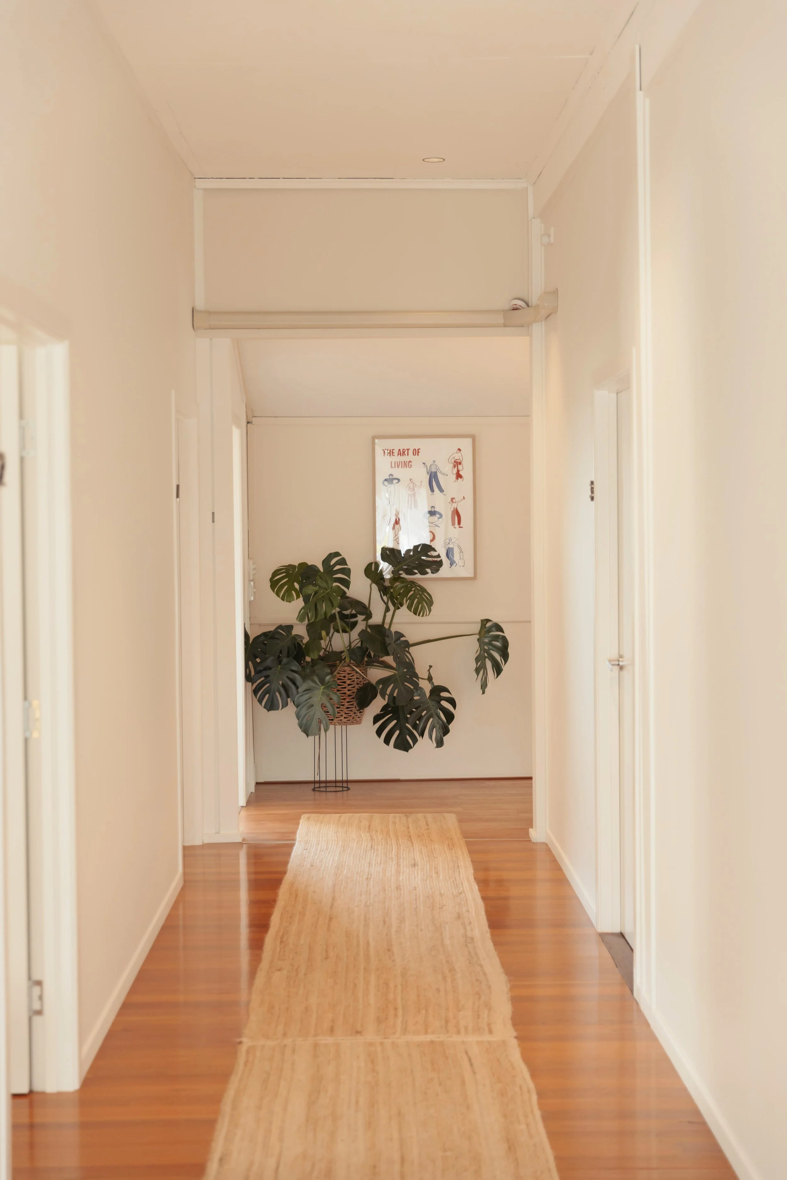 Therapy clinic hallway with light-colored walls and wooden flooring, a seagrass runner rug, two closed doors on each side, a potted monstera plant at the end of the hallway, and artwork titled 'The Art of Living' hanging on the wall behind the plant.