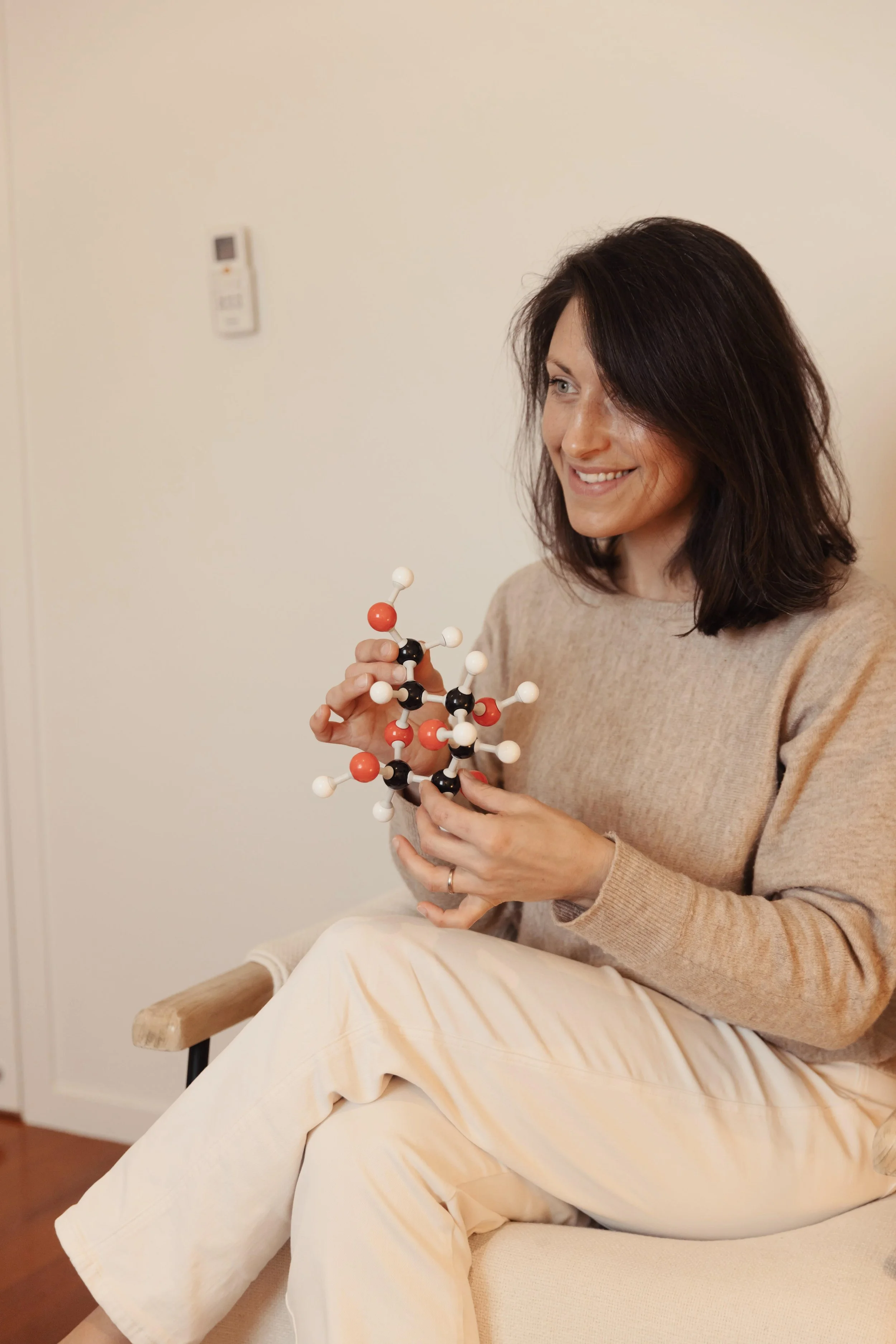 Somatic nutrition therapist and dietitian Emily Hahn sitting on a therapy chair holding a molecular model of glucose, with black, white, and red spheres, smiling warmly at the person she is looking at.