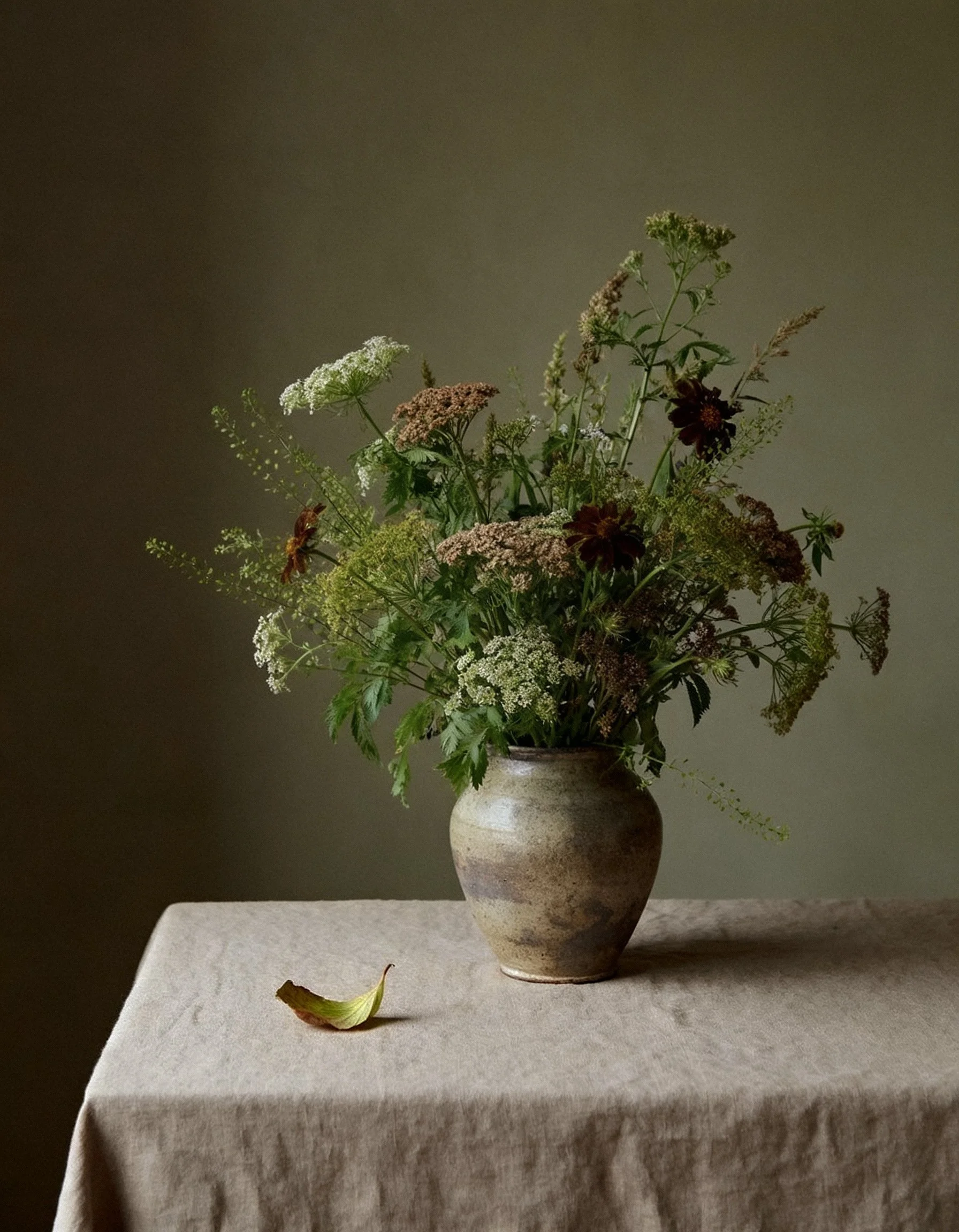 A rustic ceramic vase filled with assorted wildflowers on a beige cloth-covered table, with a grayish-green background and a single fallen leaf.