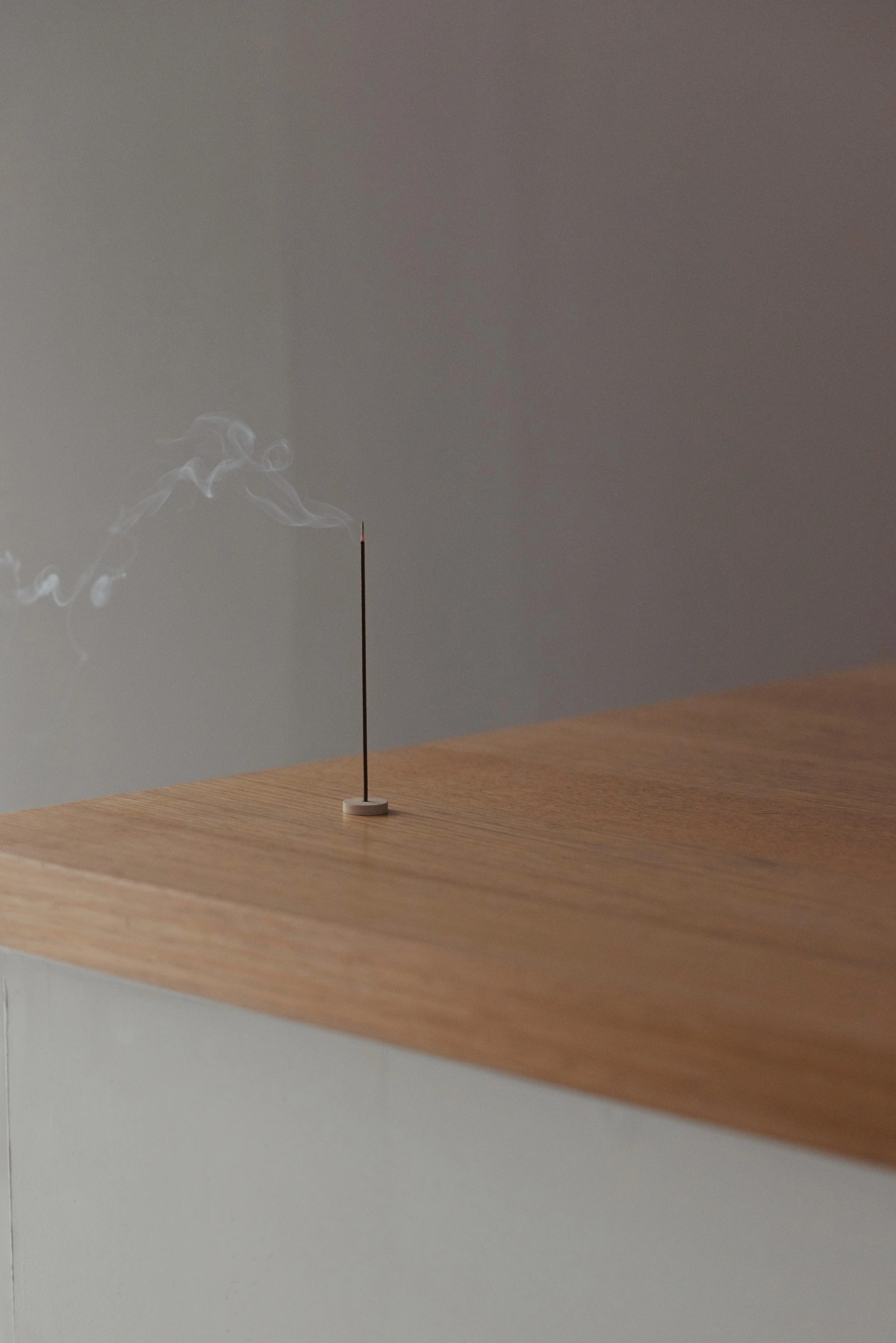 A white burning incense stick with smoke curling to the left on a wooden surface in front of a grey wall.