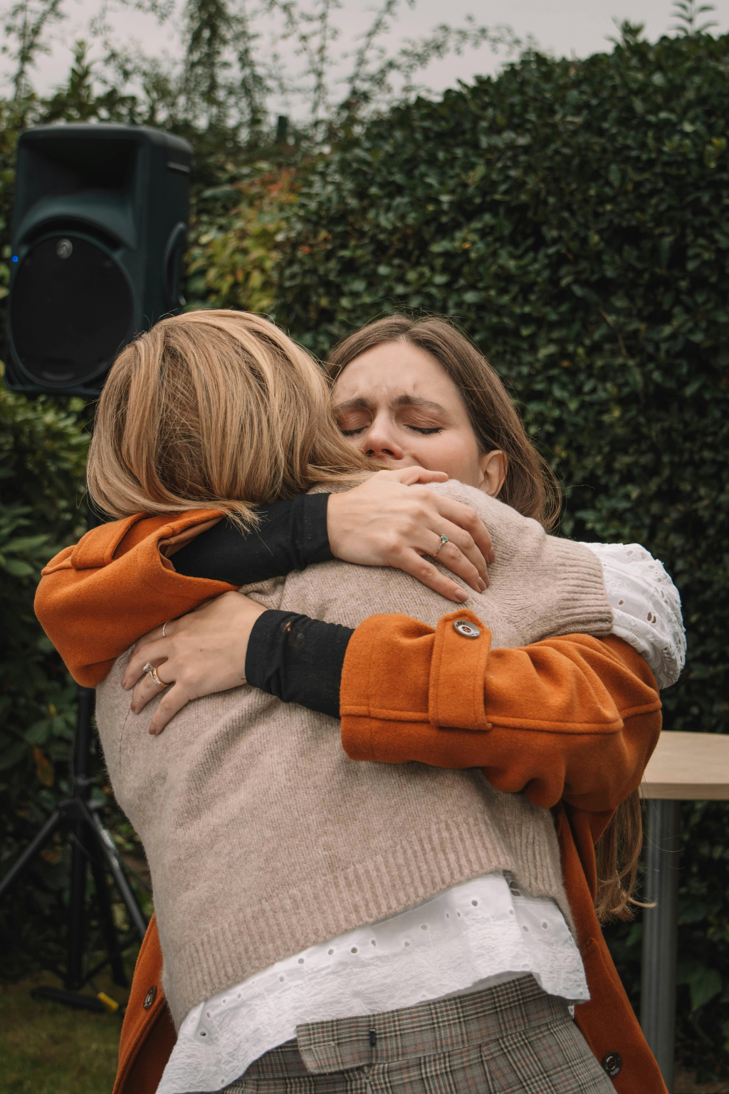 Two women hugging each other warmly outdoors, surrounded by greenery. There is an expression of relief on one woman's face as she is held.