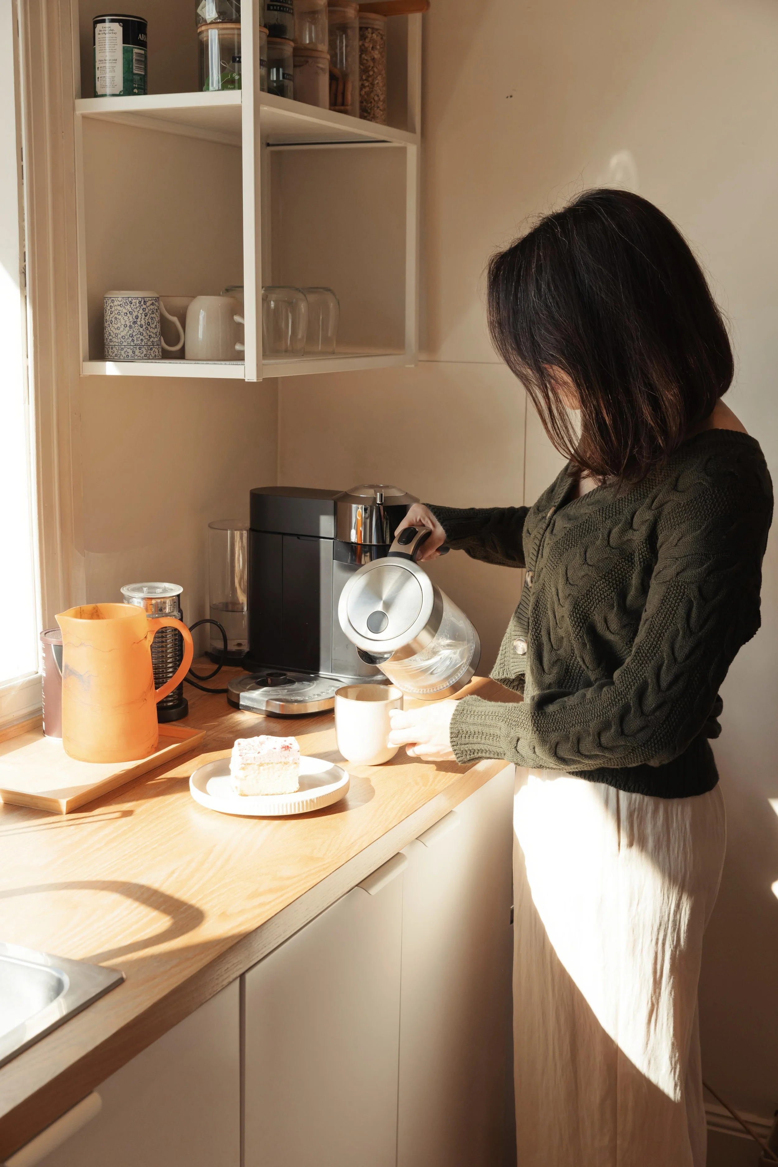 A woman pouring hot water into a white mug in a cozy kitchen with a wooden countertop, a coffee maker, and a slice of layered cake on a plate.