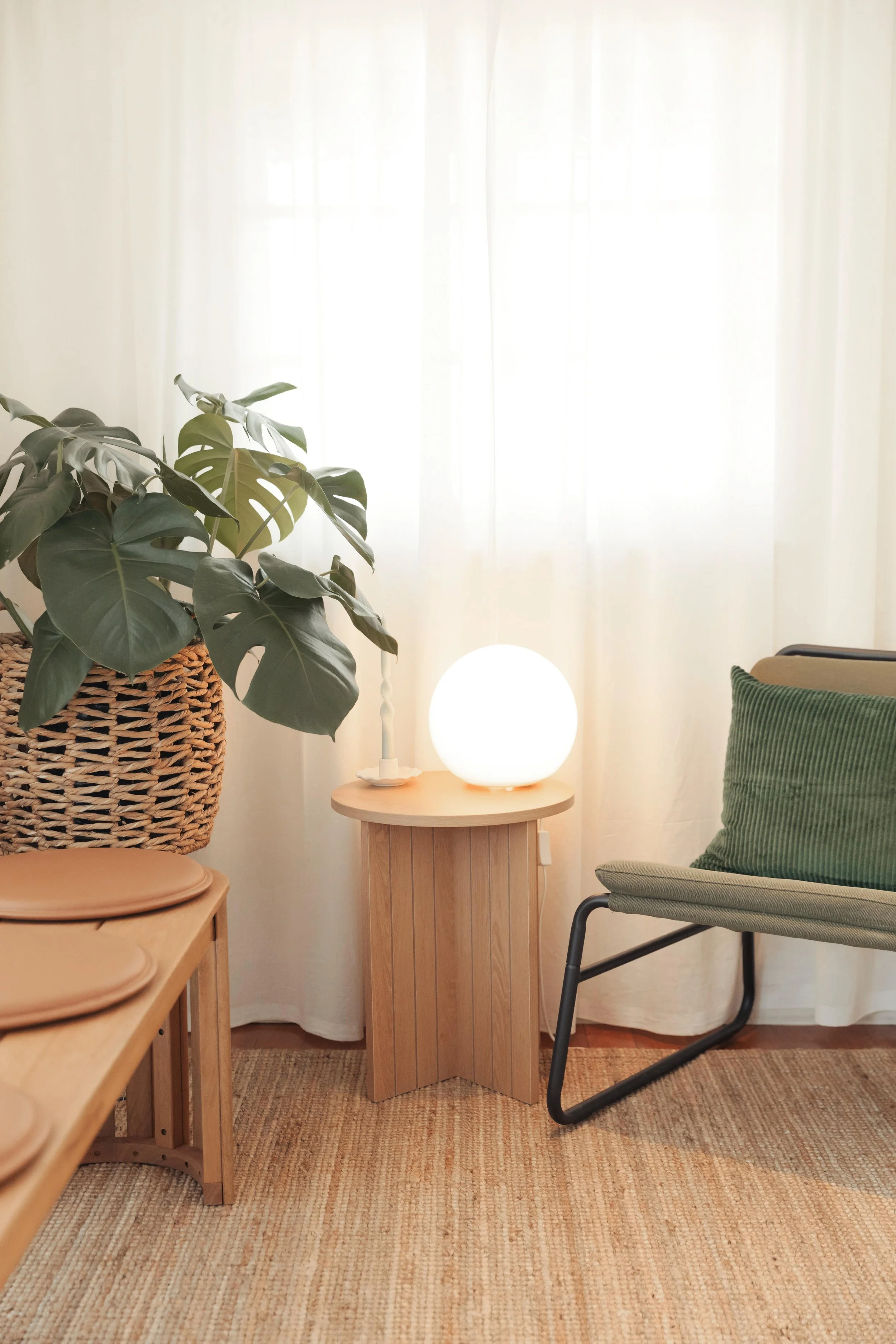 An inviting therapy clinic waiting room with a large potted monstera plant on a wicker basket, a small wooden side table with a glowing round lamp, a green cushion on a modern chair, and beige curtains in the background.