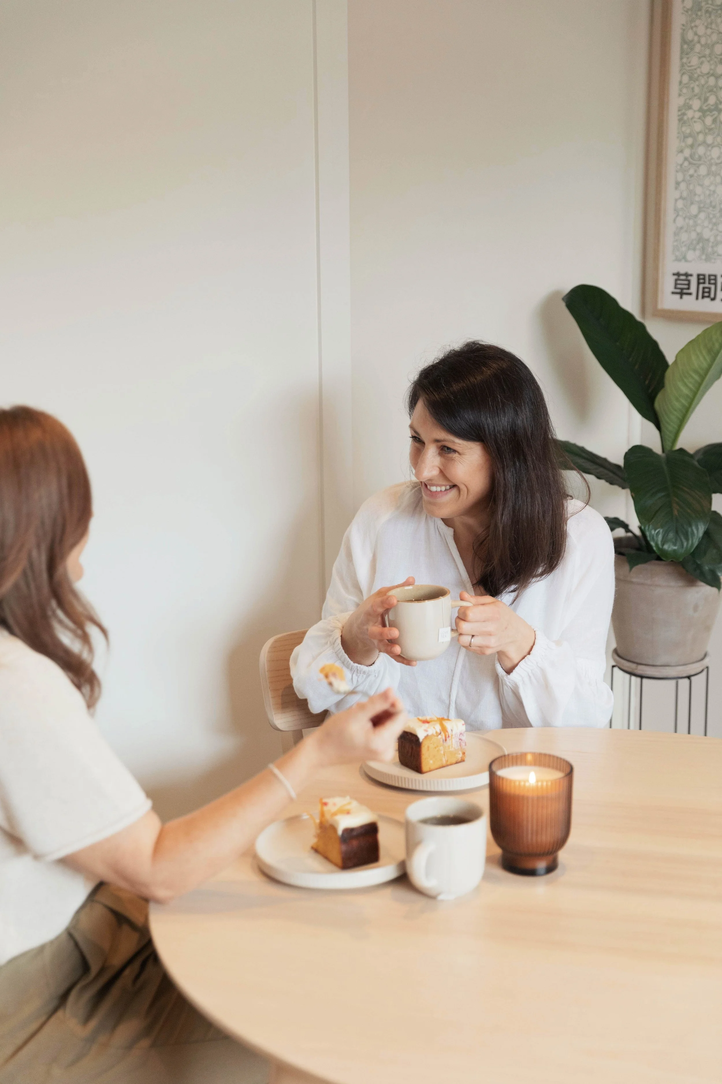 Somatic nutrition therapist and dietitian Emily Hahn enjoying a cup of tea and cake at a round table with a second woman, in a cozy, well-lit room with a potted plant in the background.