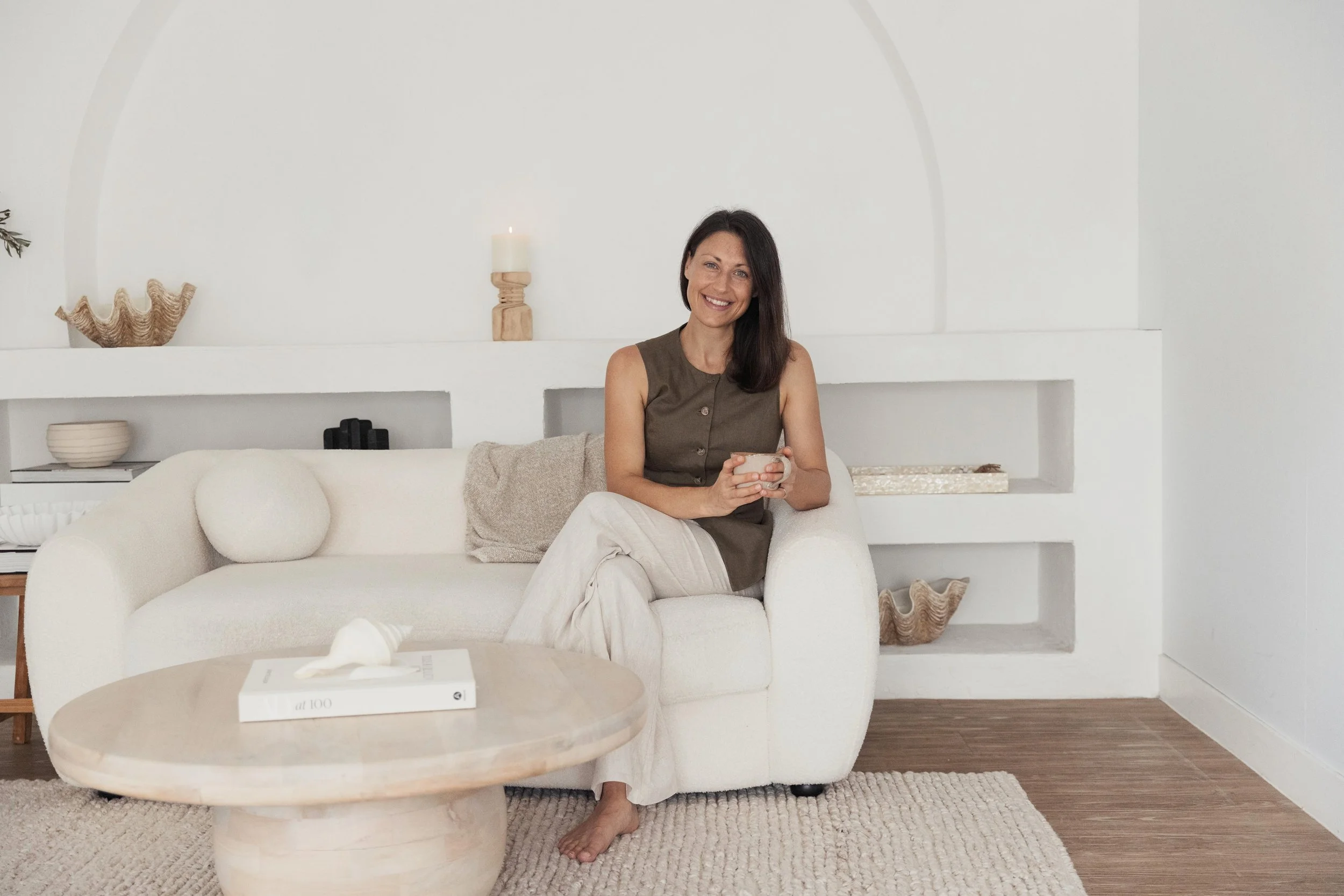 Dietitian Emily Hahn sitting on a white sofa, smiling and holding a mug. The room has a minimalist, neutral-toned decor with a wooden coffee table, decorative shells, a white book, and a shelf built into the white wall behind her.