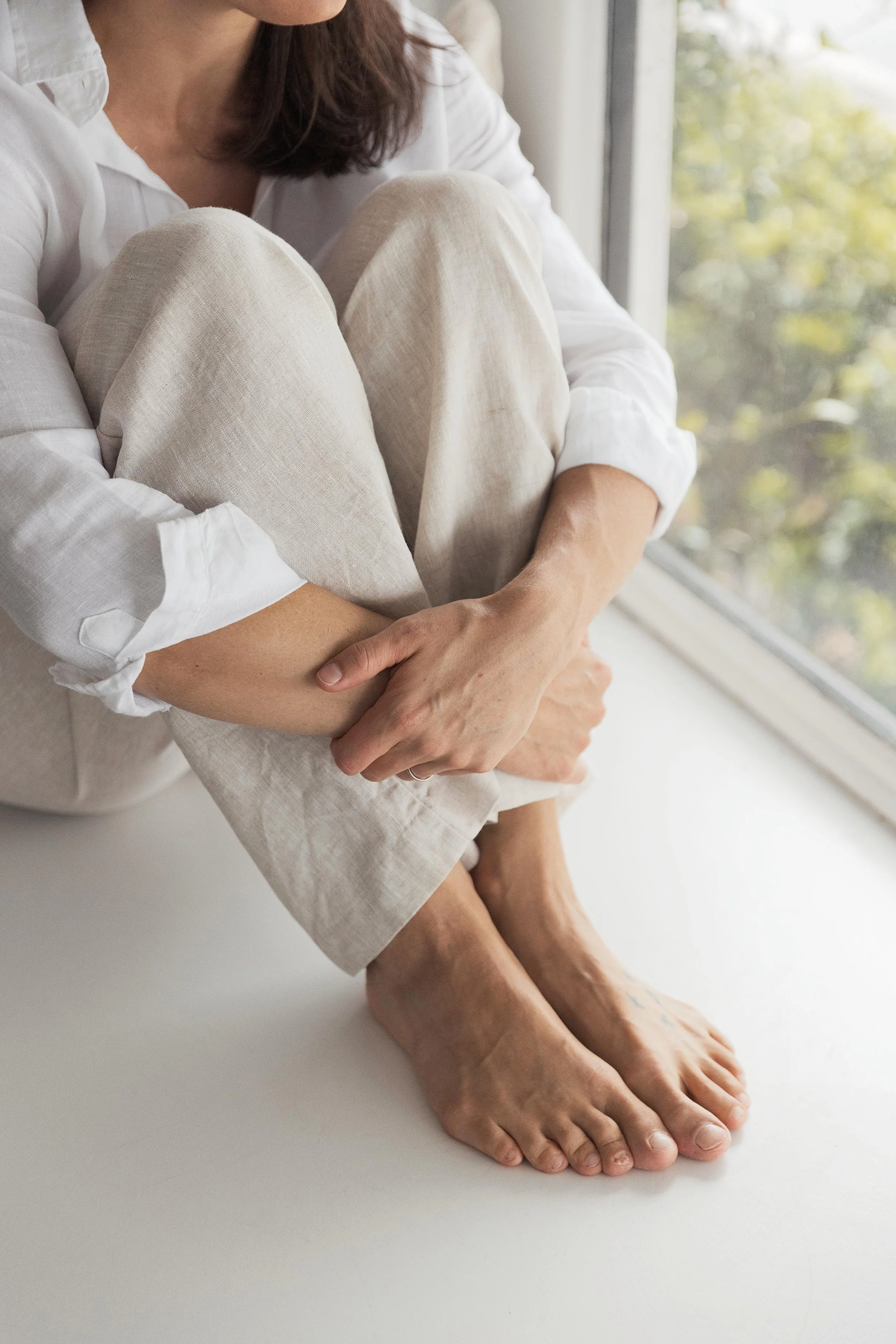 A woman sits with her knees drawn up to her chest by a window, hugging her legs with her arms, wearing beige linen pants and a white shirt, with visible bare feet.