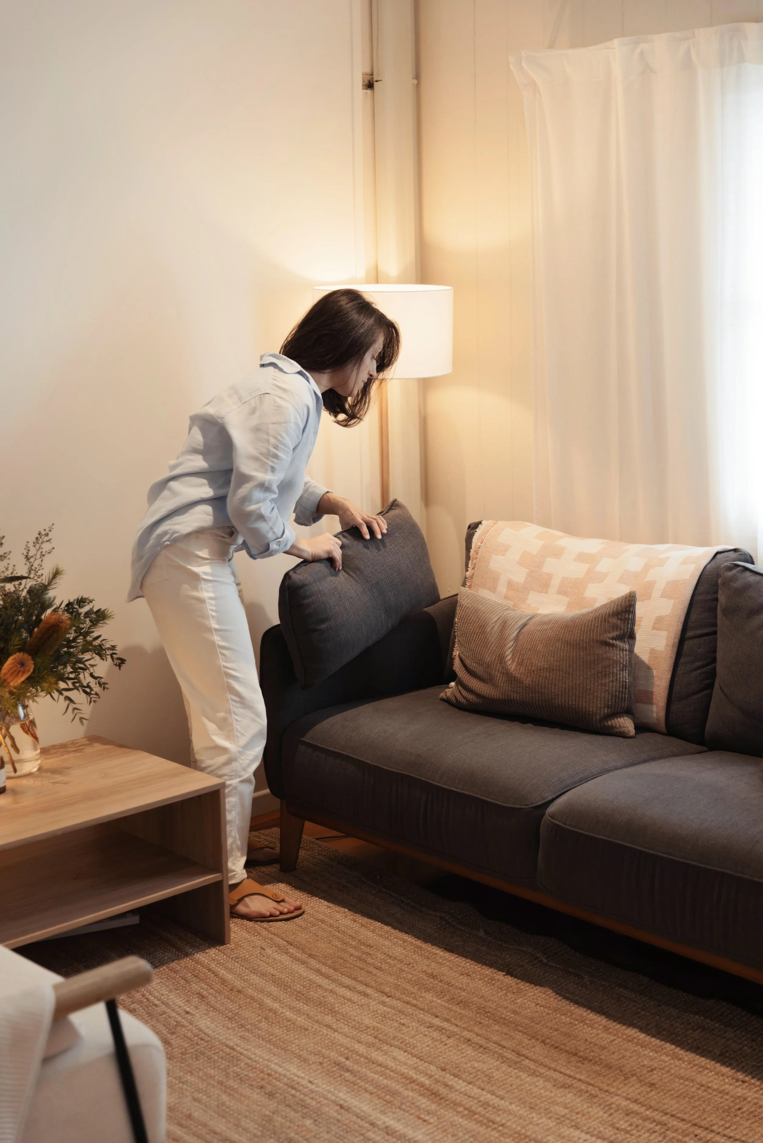 Somatic nutrition therapist and dietitian Emily Hahn in white trousers and a sky-blue linen shirt flipping a cushion on a dark gray sofa in a cozy therapy room with a wooden side table, a flower vase, a lamp, and cream-colored curtains.