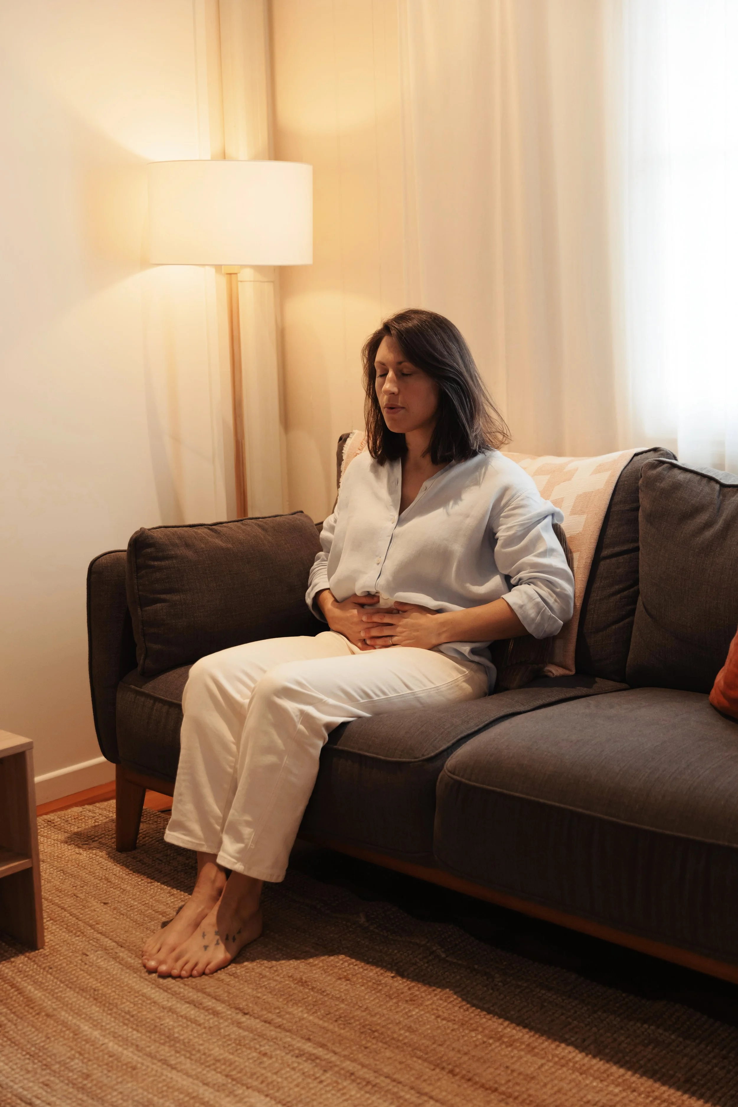 A woman sitting on a dark gray sofa in a warmly lit therapy room, holding her stomach with both hands, eyes closed, wearing a blue shirt and white pants, with beige curtains and a window behind her.