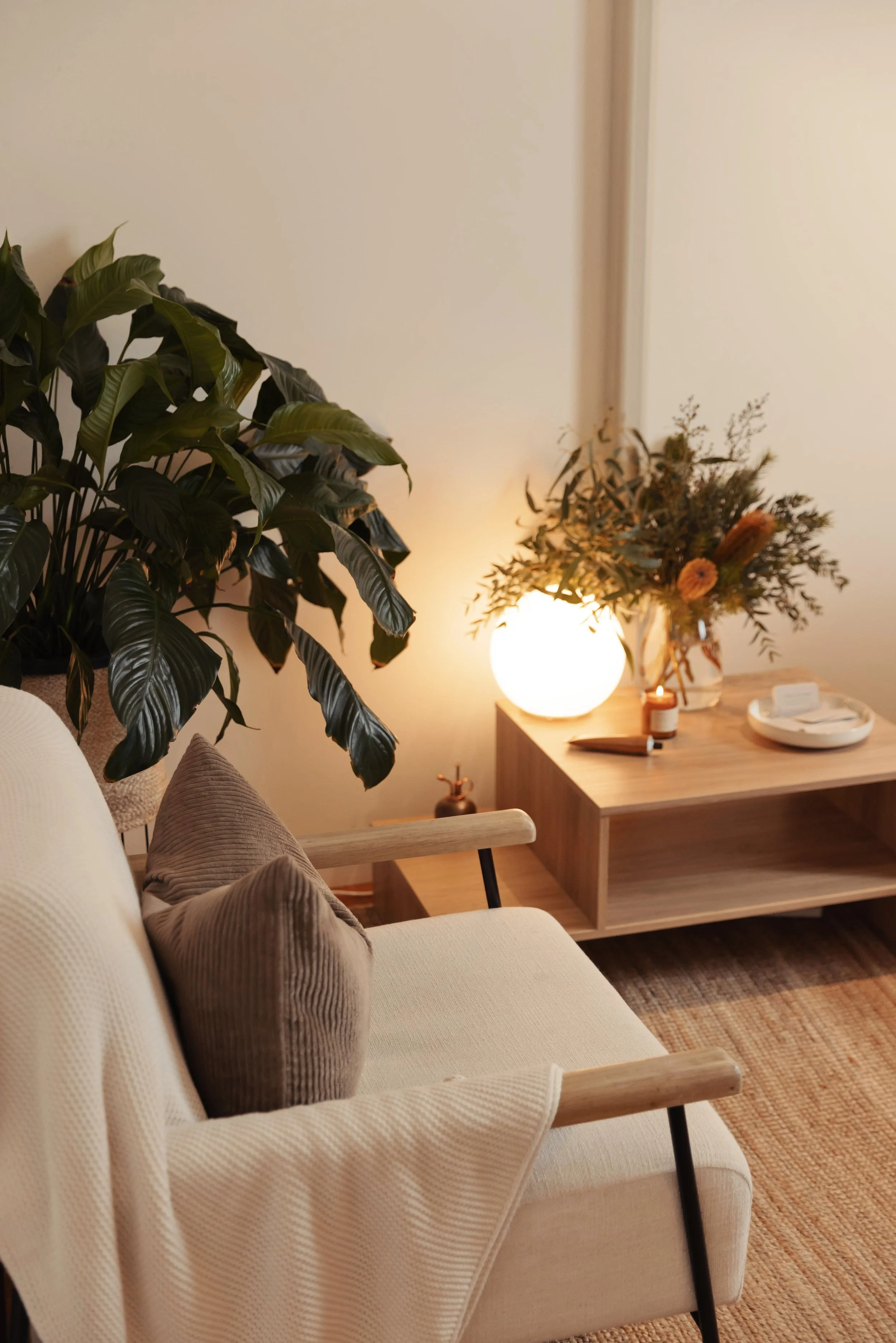 Cozy therapy room corner with a cream-white therapist chair, brown pillows, a large green plant, and a wooden side table with a glowing round lamp, flower arrangement, and small lit candle in a brown jar.