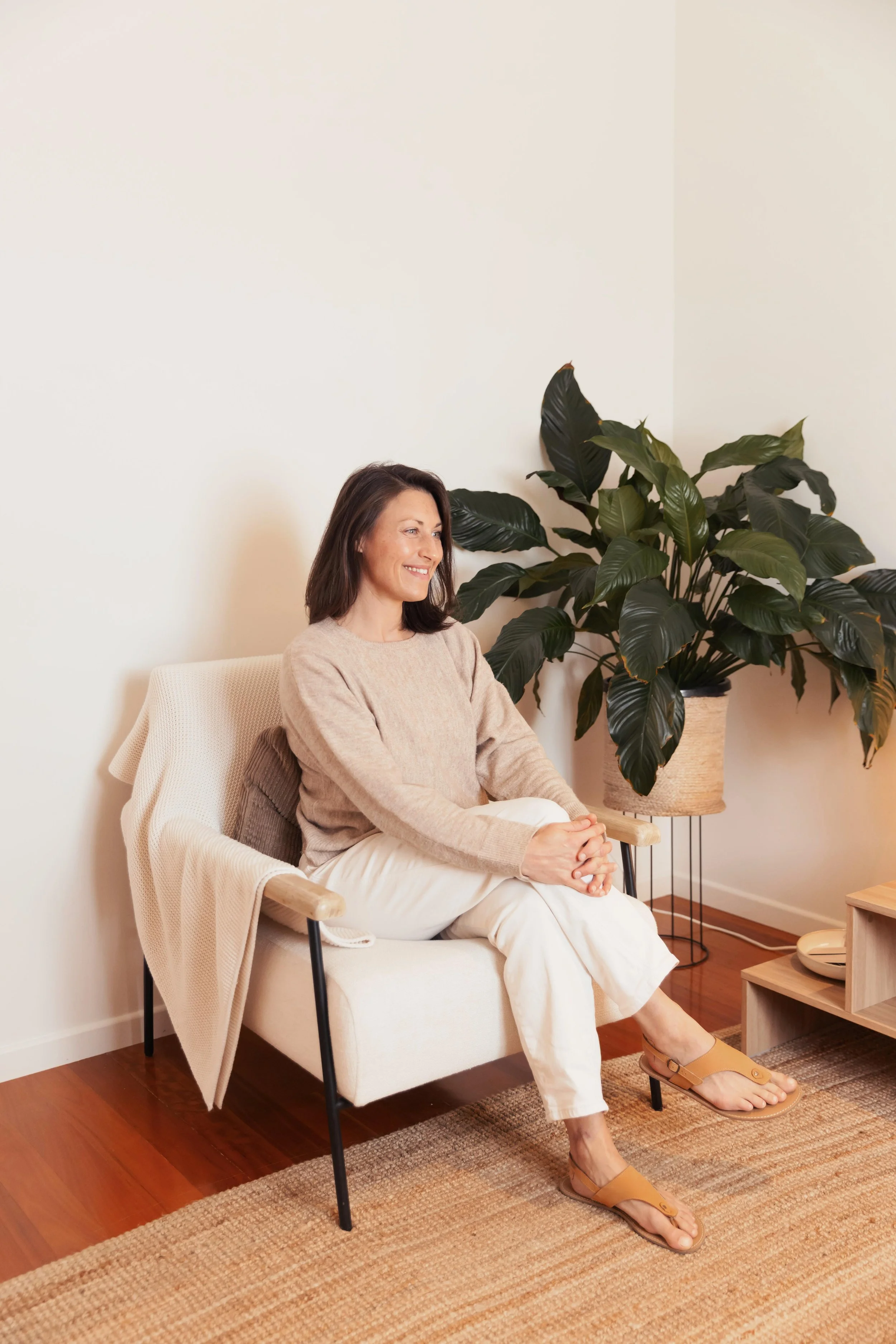 Somatic nutrition therapist and dietitian Emily Hahn sitting on a cream-colored armchair in a therapy room, smiling, with a large potted plant beside her.