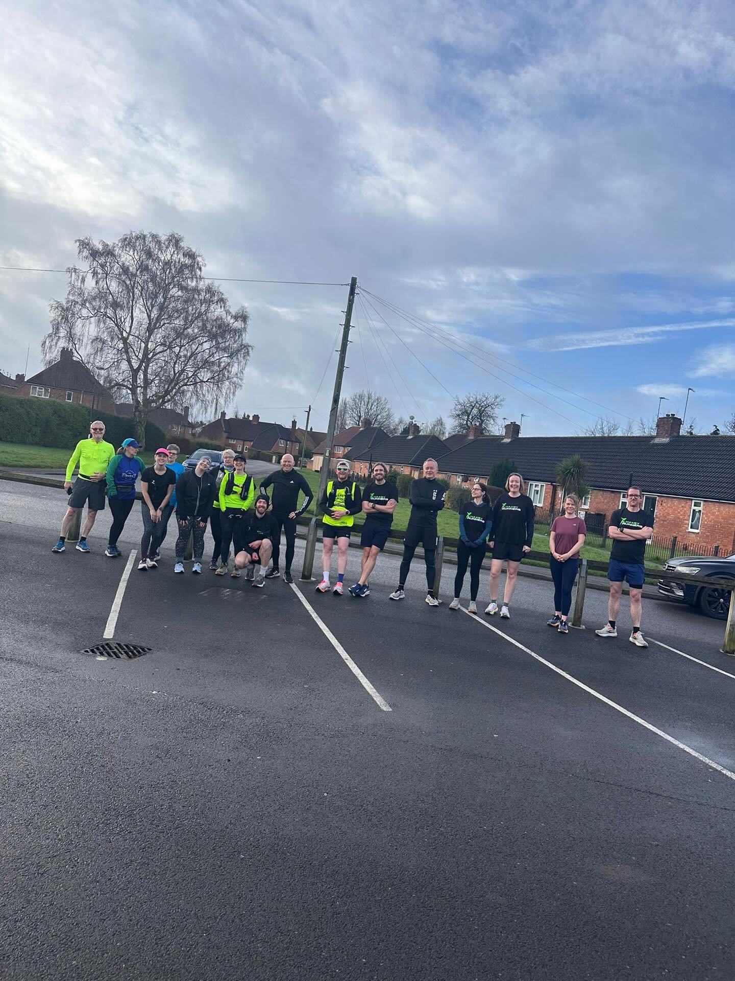 Group of people dressed in athletic clothing standing in a parking lot with houses and trees in the background on a partly cloudy day.