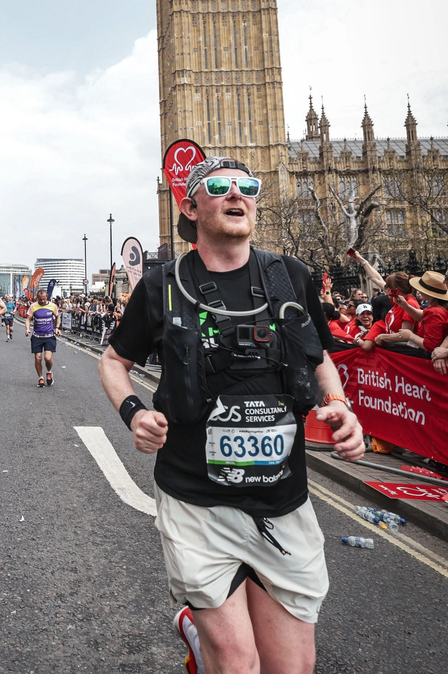 A man running in a marathon in front of Big Ben in London, UK. He is wearing sunglasses, a cap, and a race bib number 63360, with a crowd and supporters behind him.