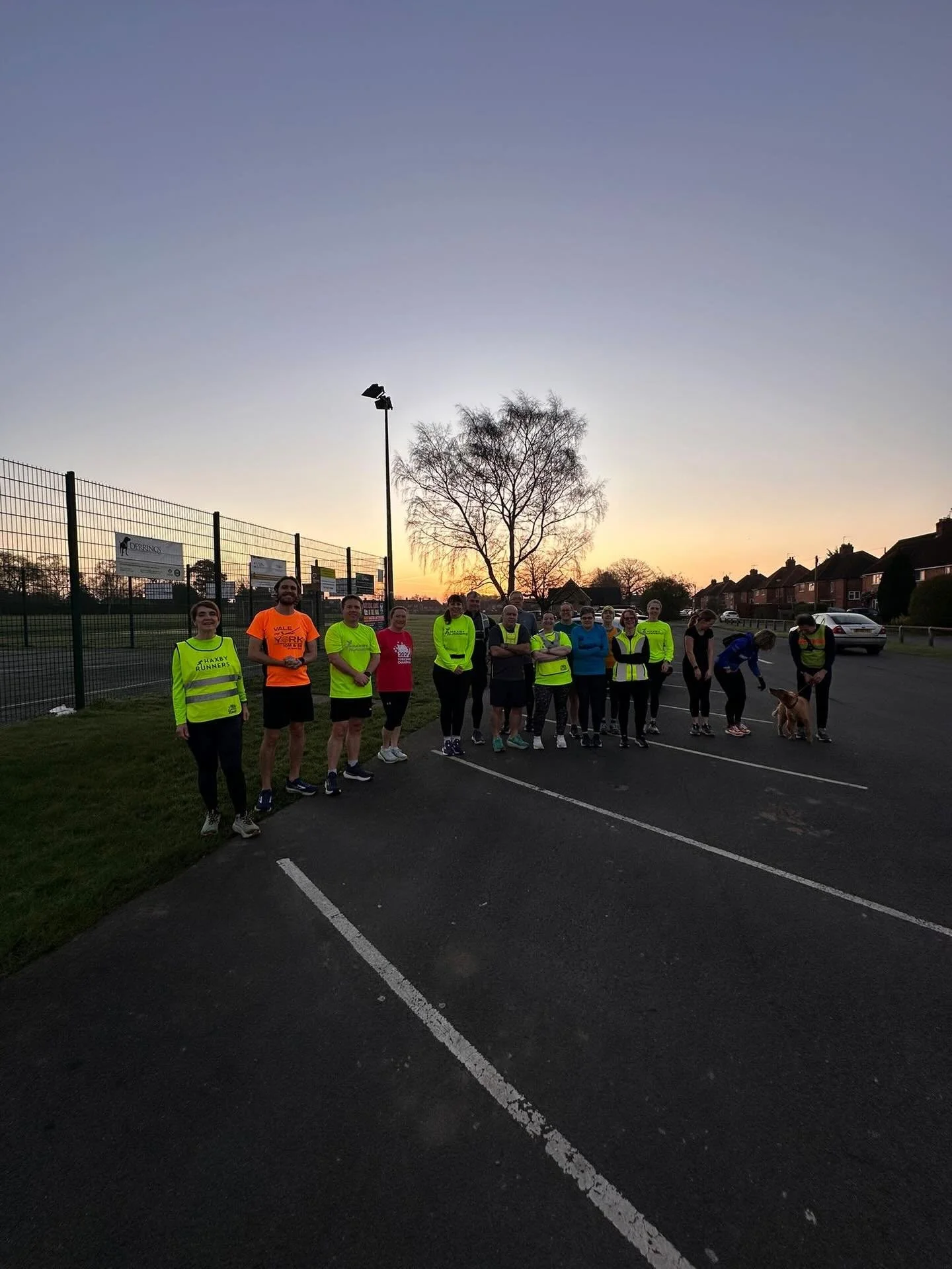 Group of people in bright athletic clothing standing on a track at sunset, some with reflective vests, with houses and trees in the background.