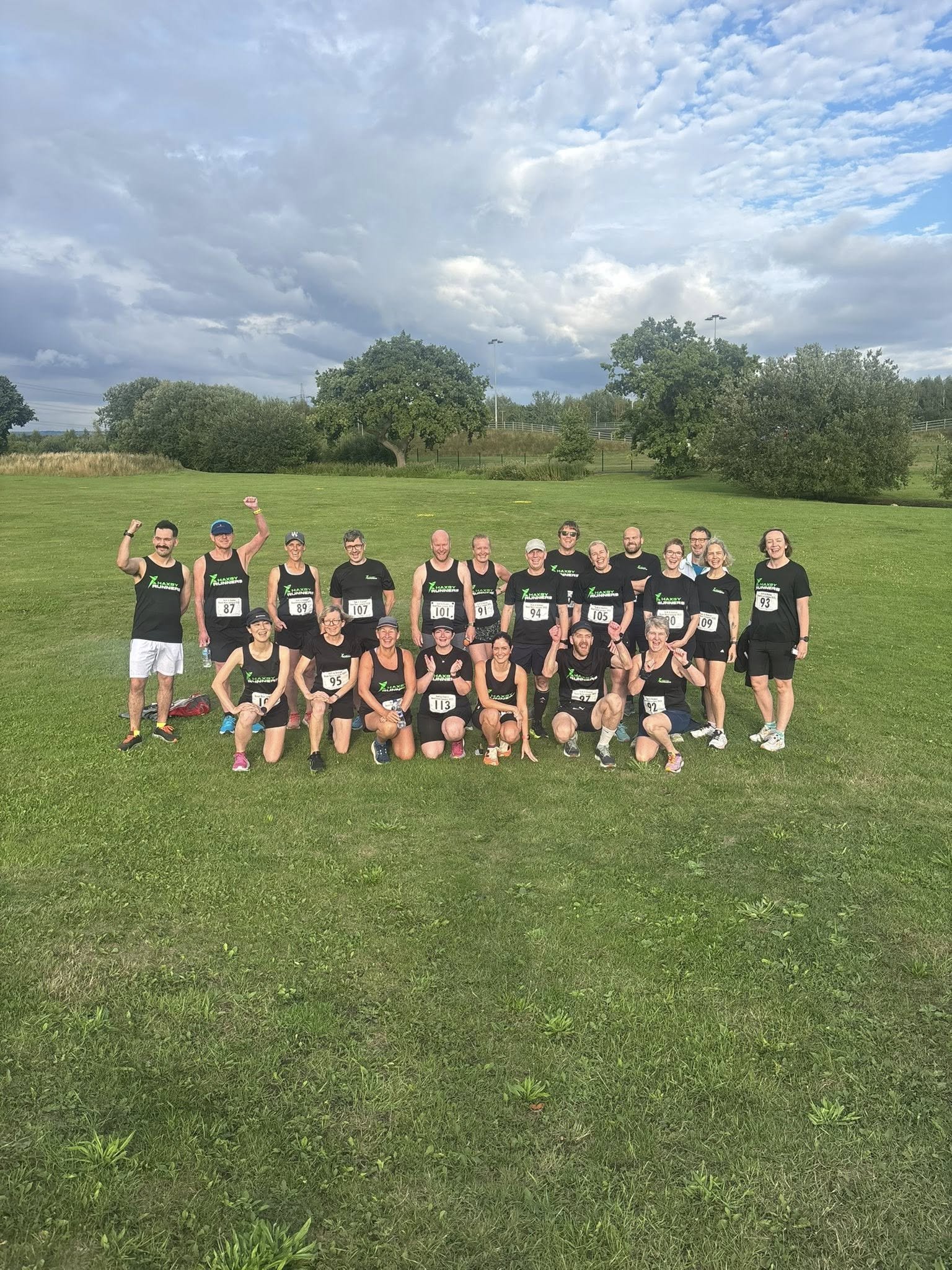 Group of people in athletic clothing standing on a grassy field, some raising their arms, with trees, a fence, and a cloudy sky in the background.