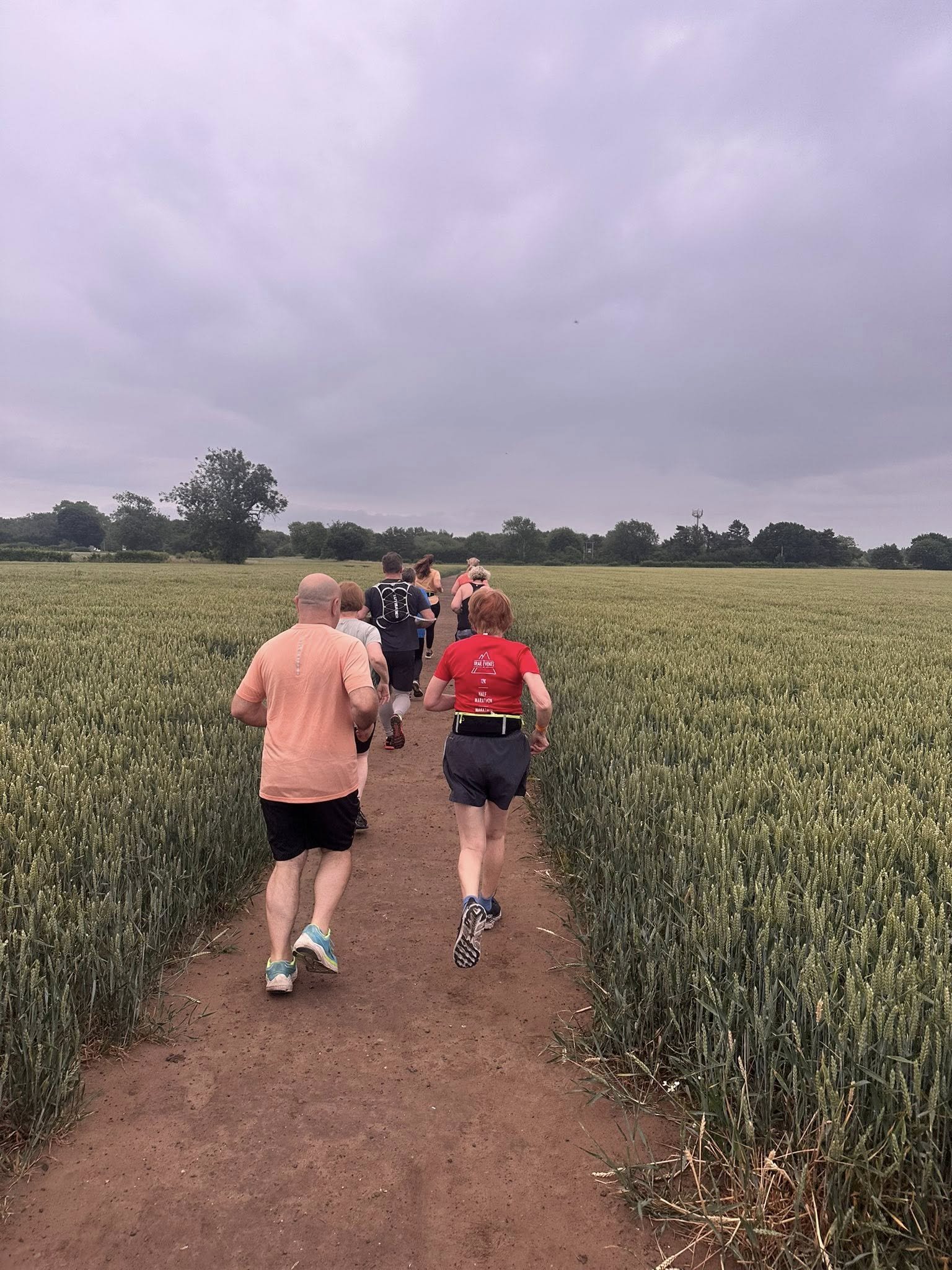 A group of people jogging on a dirt trail through a green wheat field under a cloudy sky.