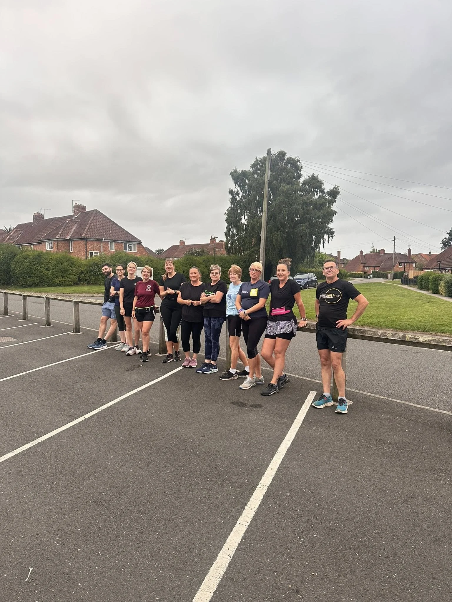Group of people dressed in athletic clothing standing in a parking lot with residential houses and trees in the background, overcast weather.