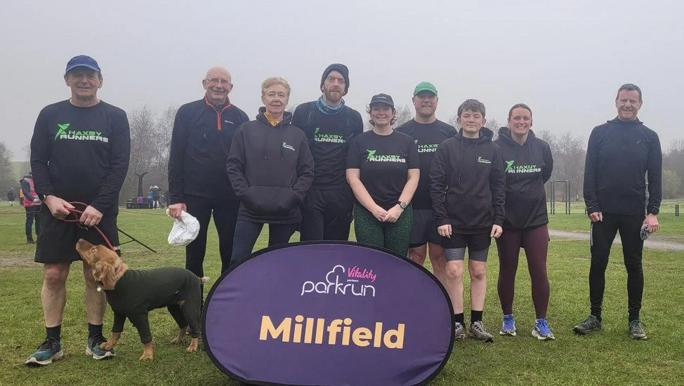 Group of nine people and one dog standing on a grassy field with a signs that says 'Millfield' and 'parkrun.' The people are dressed in athletic clothing, with some wearing 'Hackney Runners' shirts, and appear to be participating in a race or event.