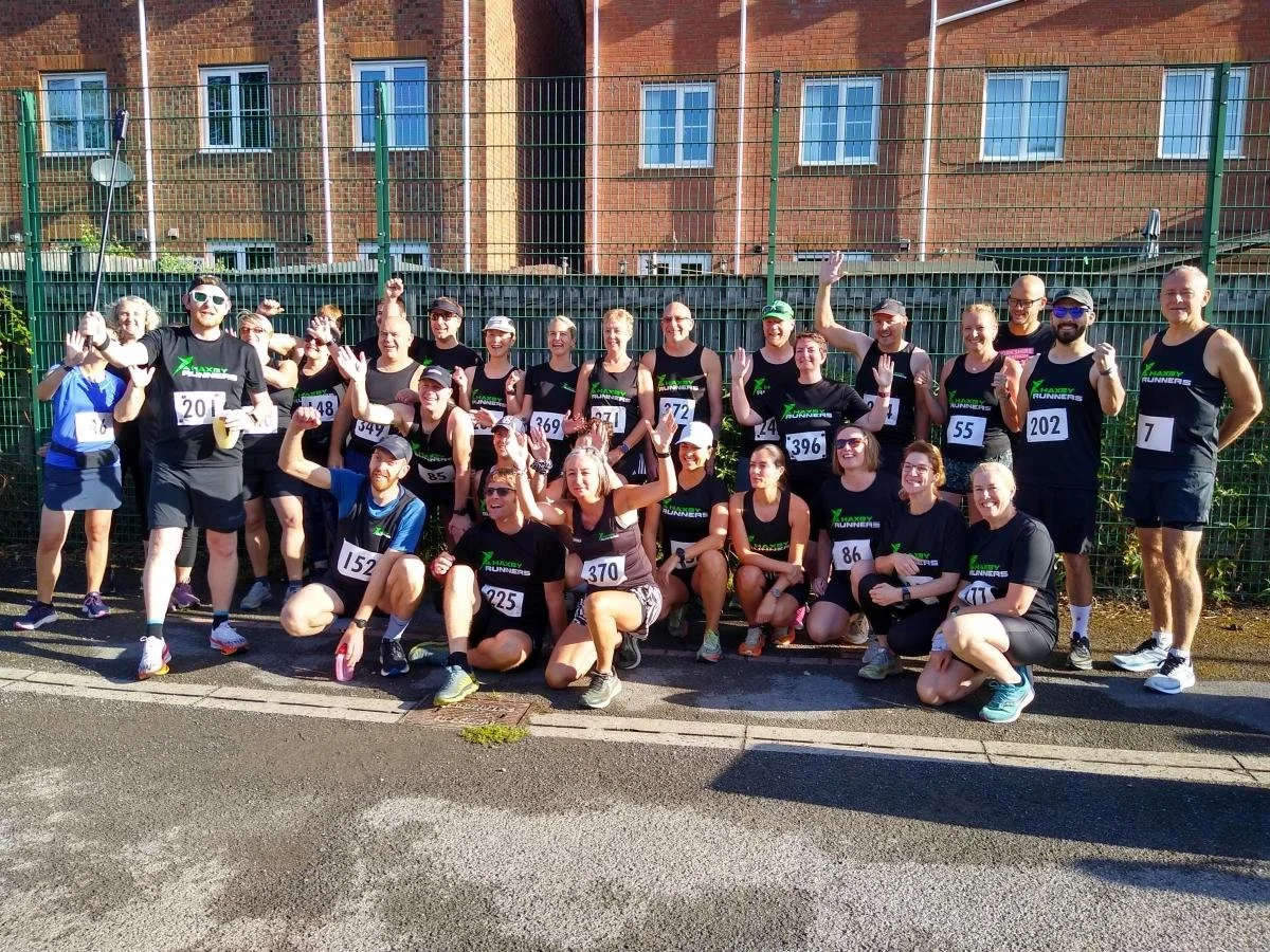 Group of runners in black athletic wear and race bibs posing and smiling outdoors after a race, with some raising their hands and making peace signs.