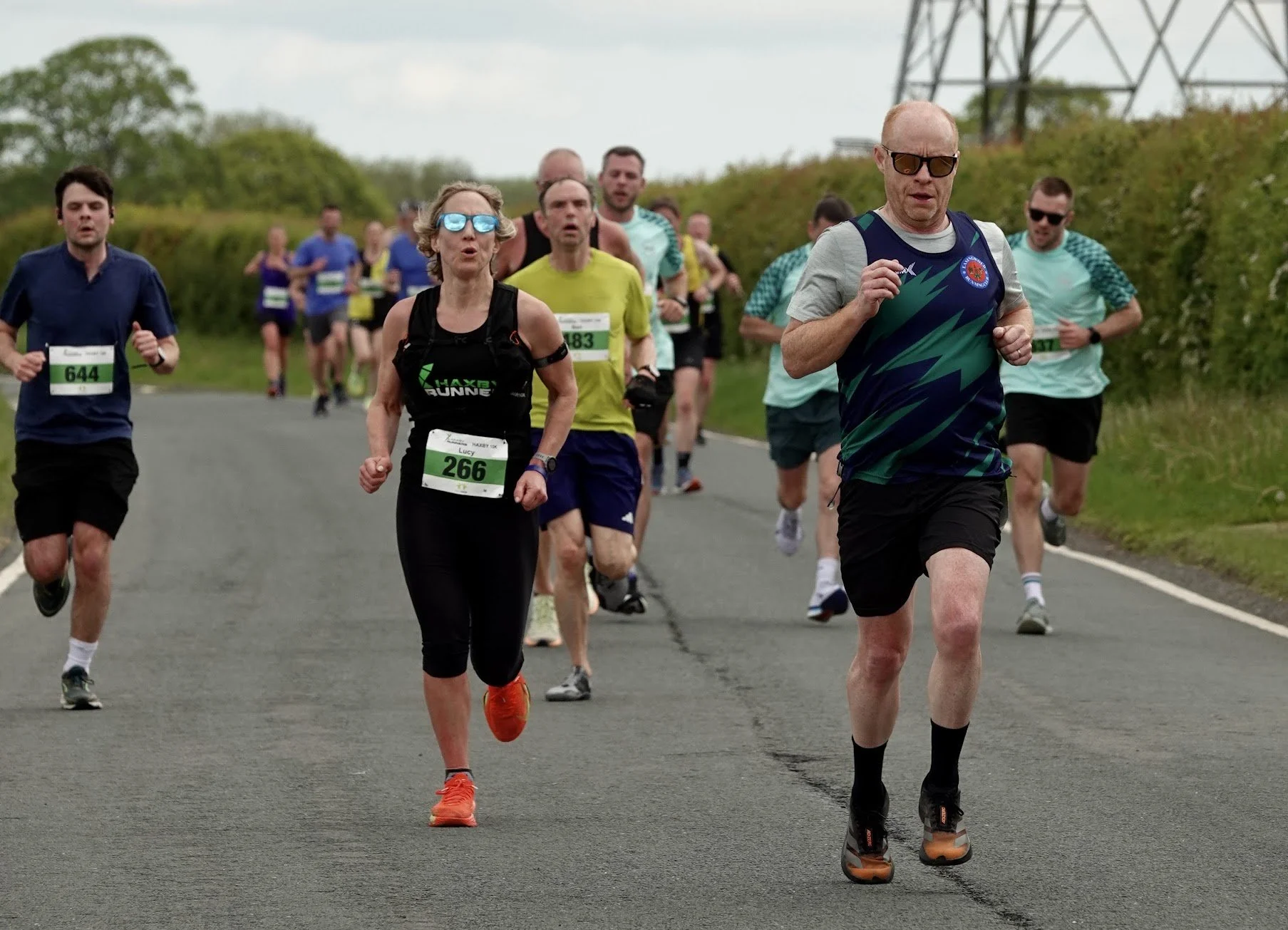 A group of people participating in a road race on a paved rural road with green bushes and trees in the background. The runners are wearing athletic clothing, numbered bibs, and sunglasses, with some showing focused expressions.