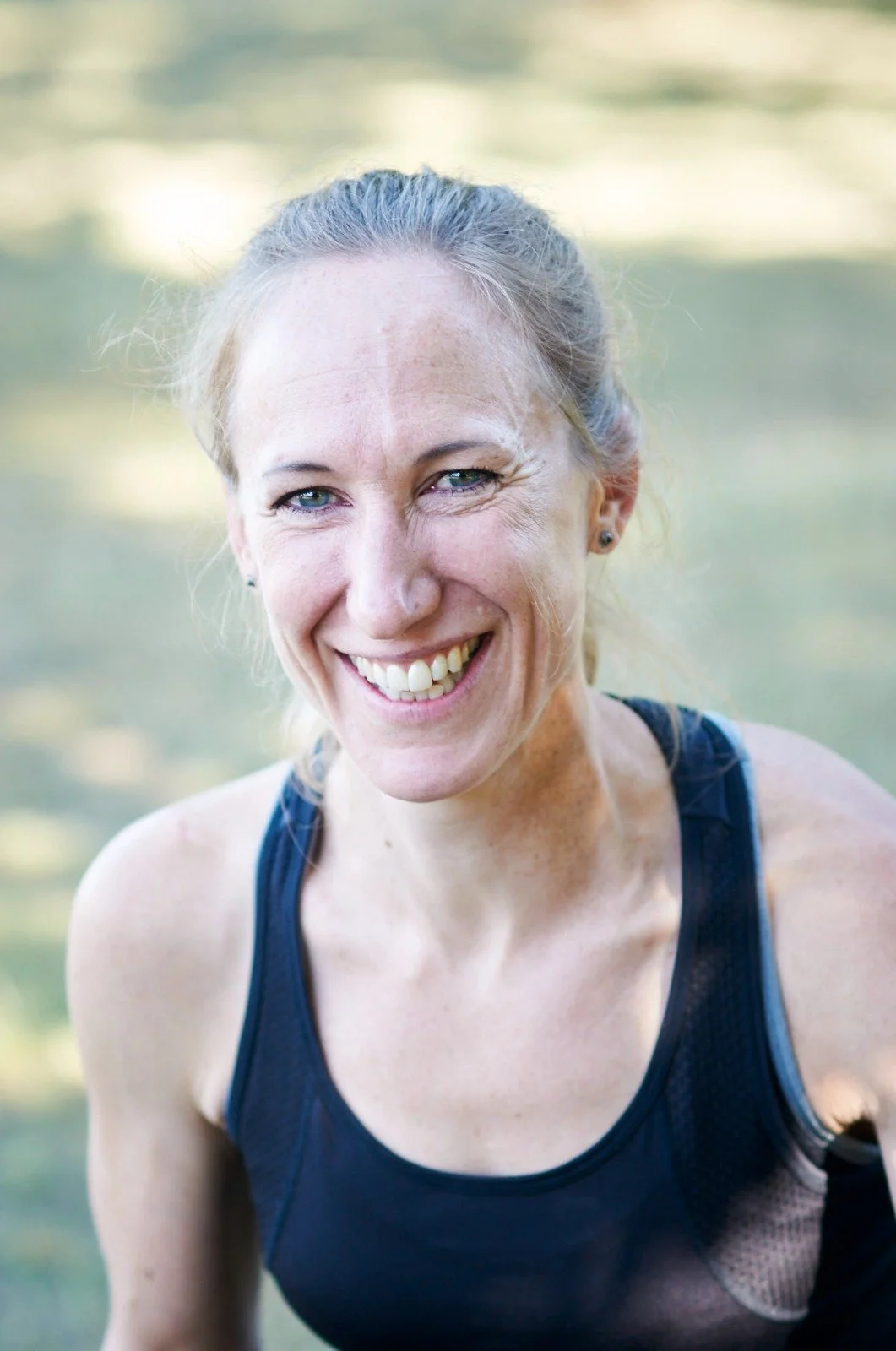 Close-up of a smiling woman with blonde hair tied back, wearing a black athletic top, outdoors in a natural setting.