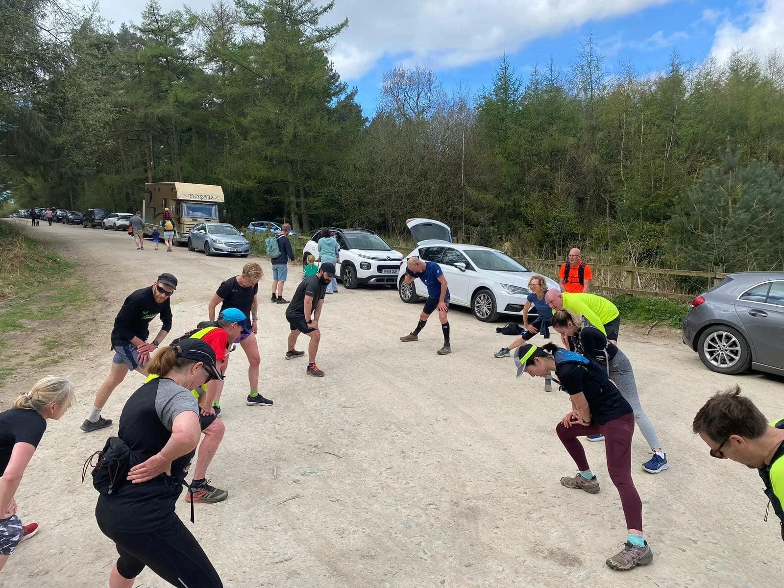 Group of people stretching outdoors on a dirt parking lot surrounded by trees, with parked cars and a blue sky with clouds in the background.