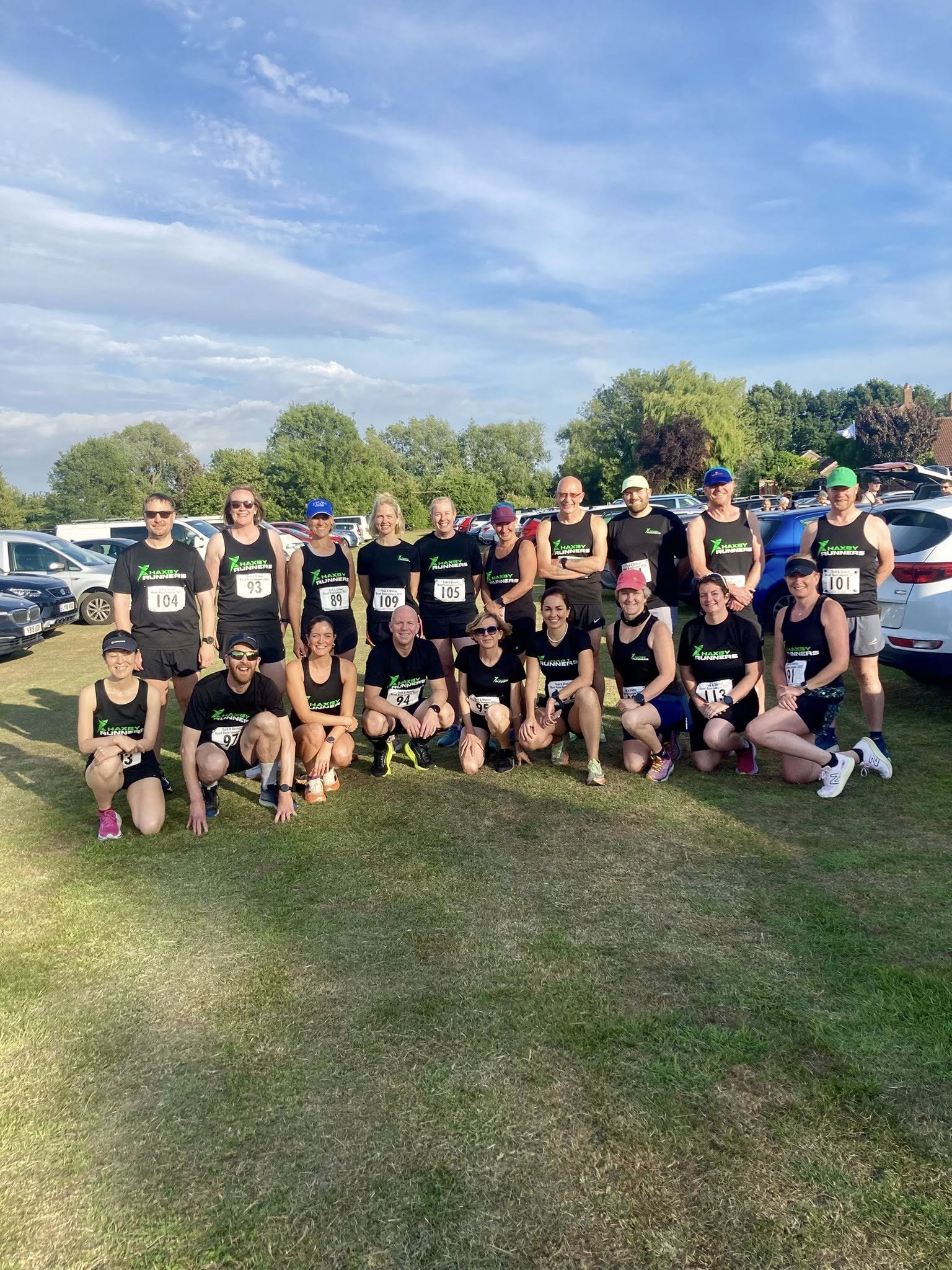 Group of runners in black athletic gear and numbered bibs posing for a photo outdoors in a grassy area with cars and trees in the background.