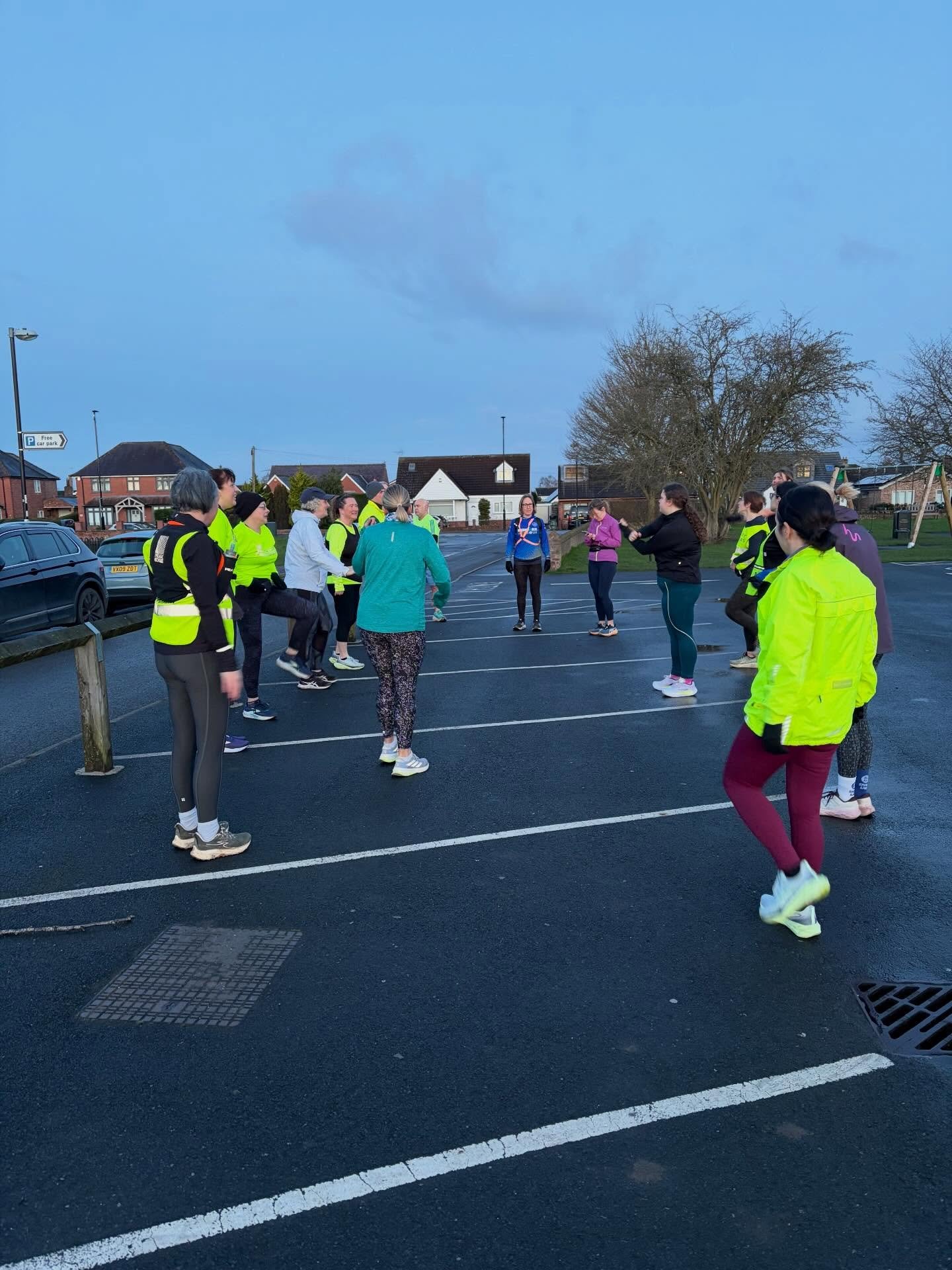 Group of people in colorful athletic clothing exercising outdoors in a parking lot during early morning or late afternoon.