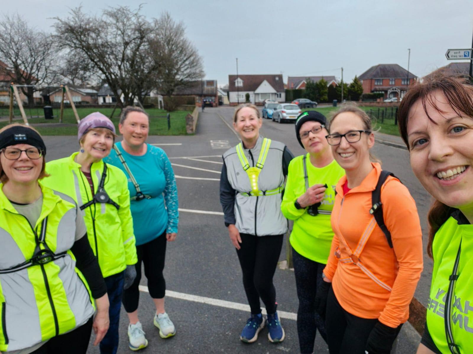 Group of seven women in running gear and reflective vests posing for a selfie in a residential neighborhood parking lot with houses and cars in the background.